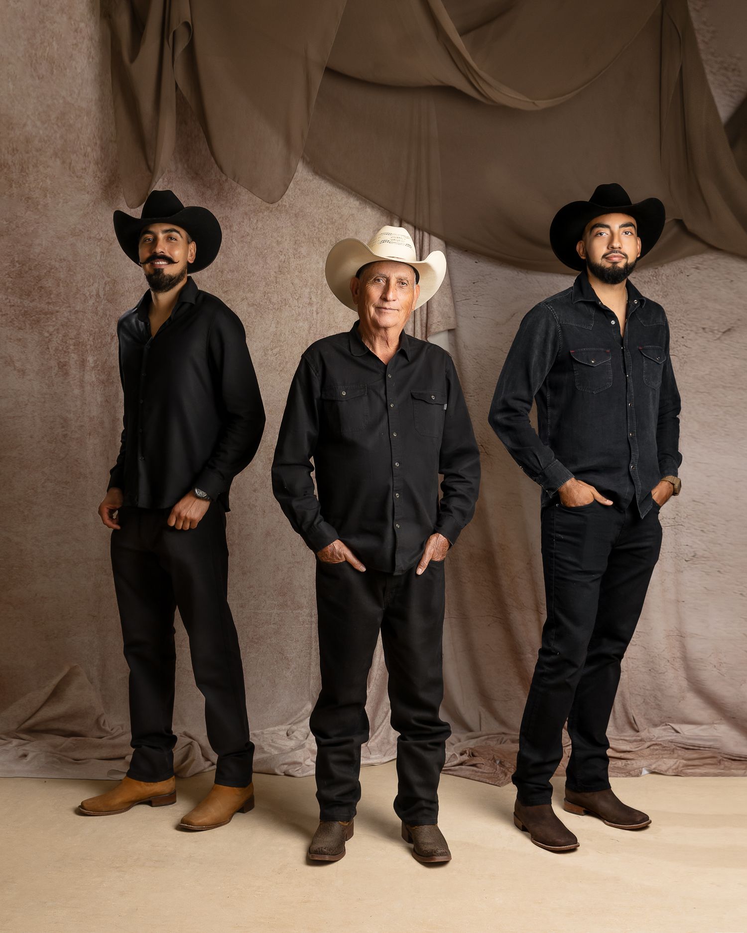 Three men in black cowboy attire pose against a draped fabric backdrop, with the center man wearing a white hat.