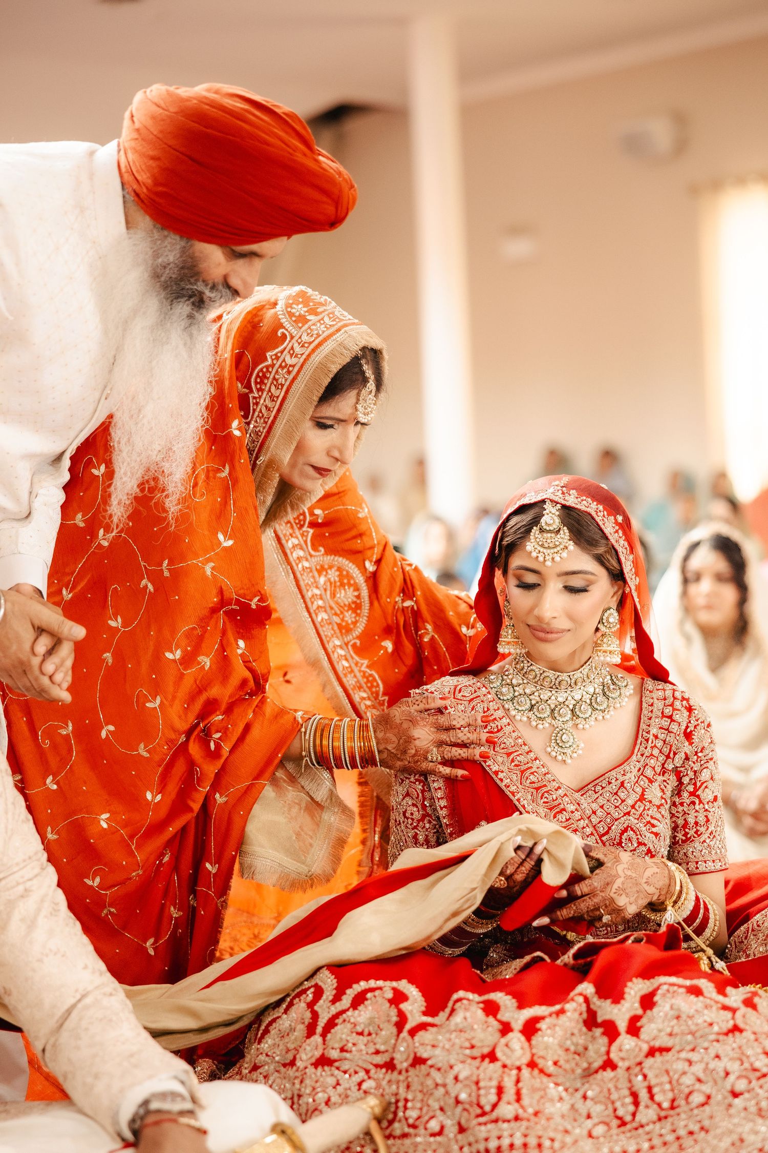 Sikh wedding ceremony with bride in red lehenga and woman in orange dupatta guided by elder in turban.