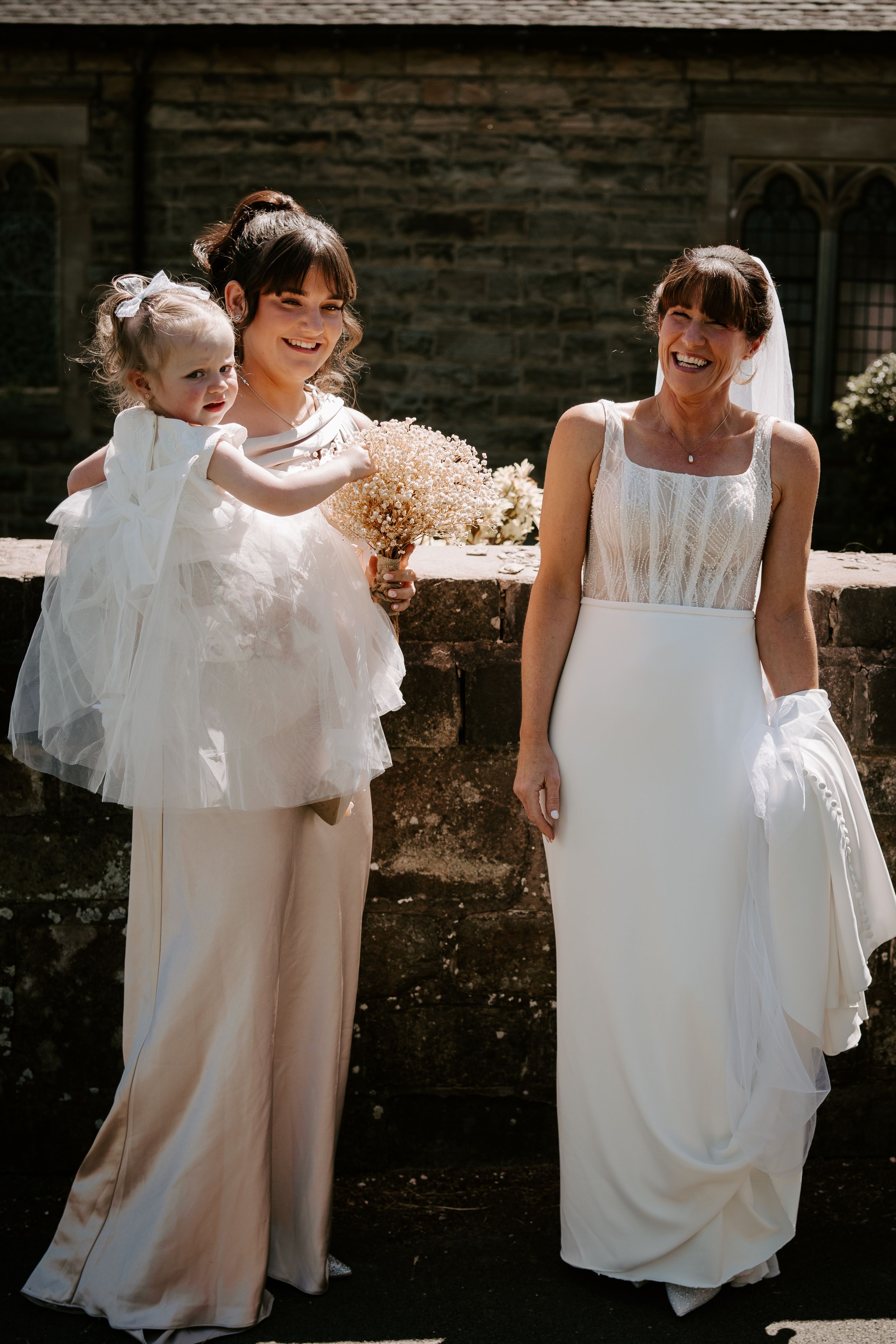 Bride in white gown laughs with bridesmaid holding a toddler in white dress outside stone church.