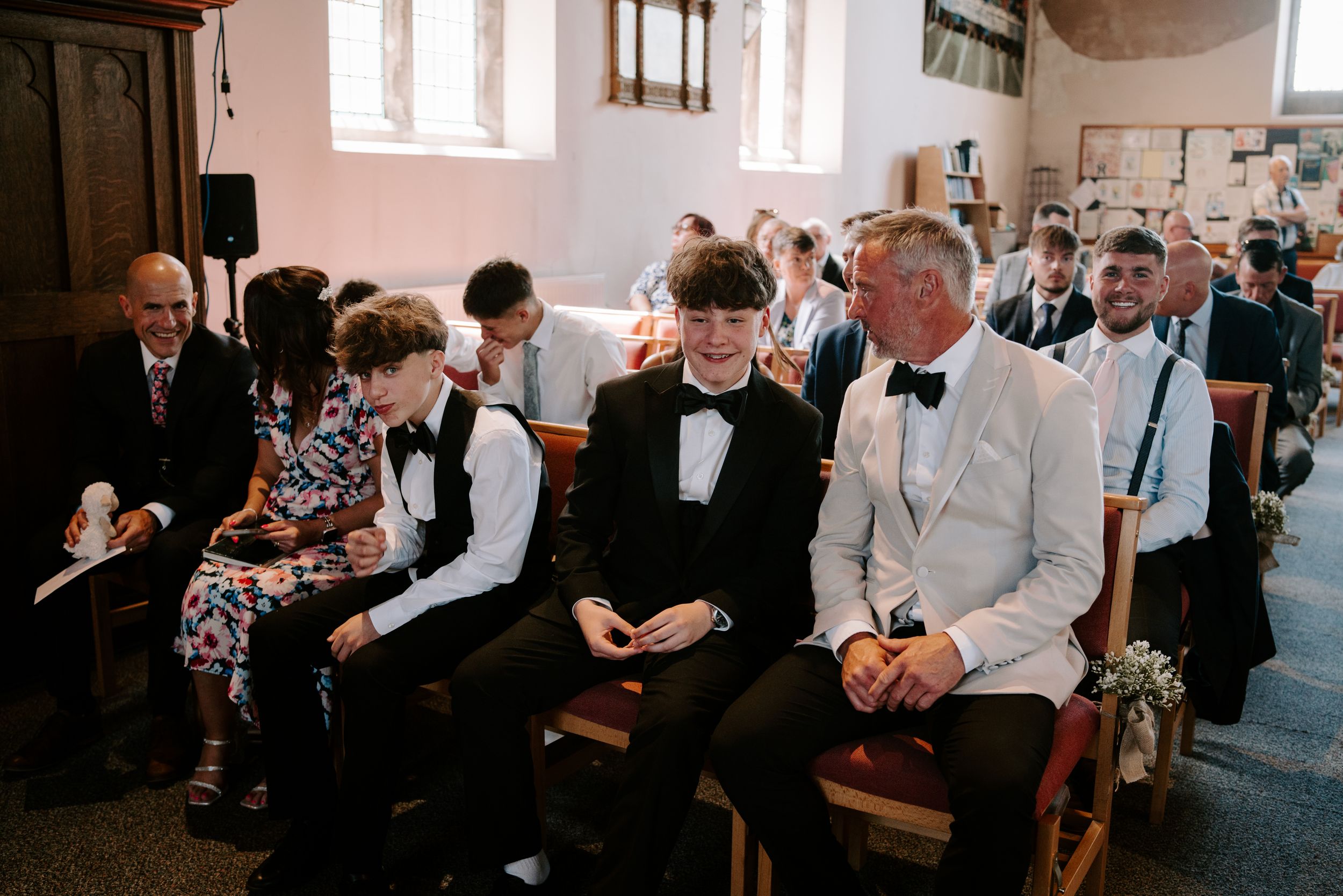 Wedding guests in formal attire smile and chat while seated in wooden church pews during ceremony.