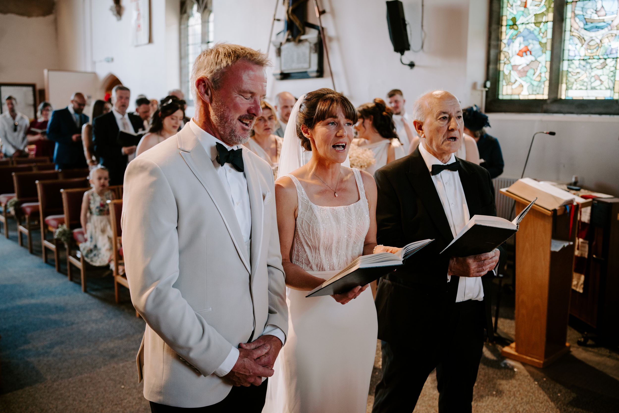 Bride and groom singing hymns with family during a church wedding ceremony, with stained glass windows in background.