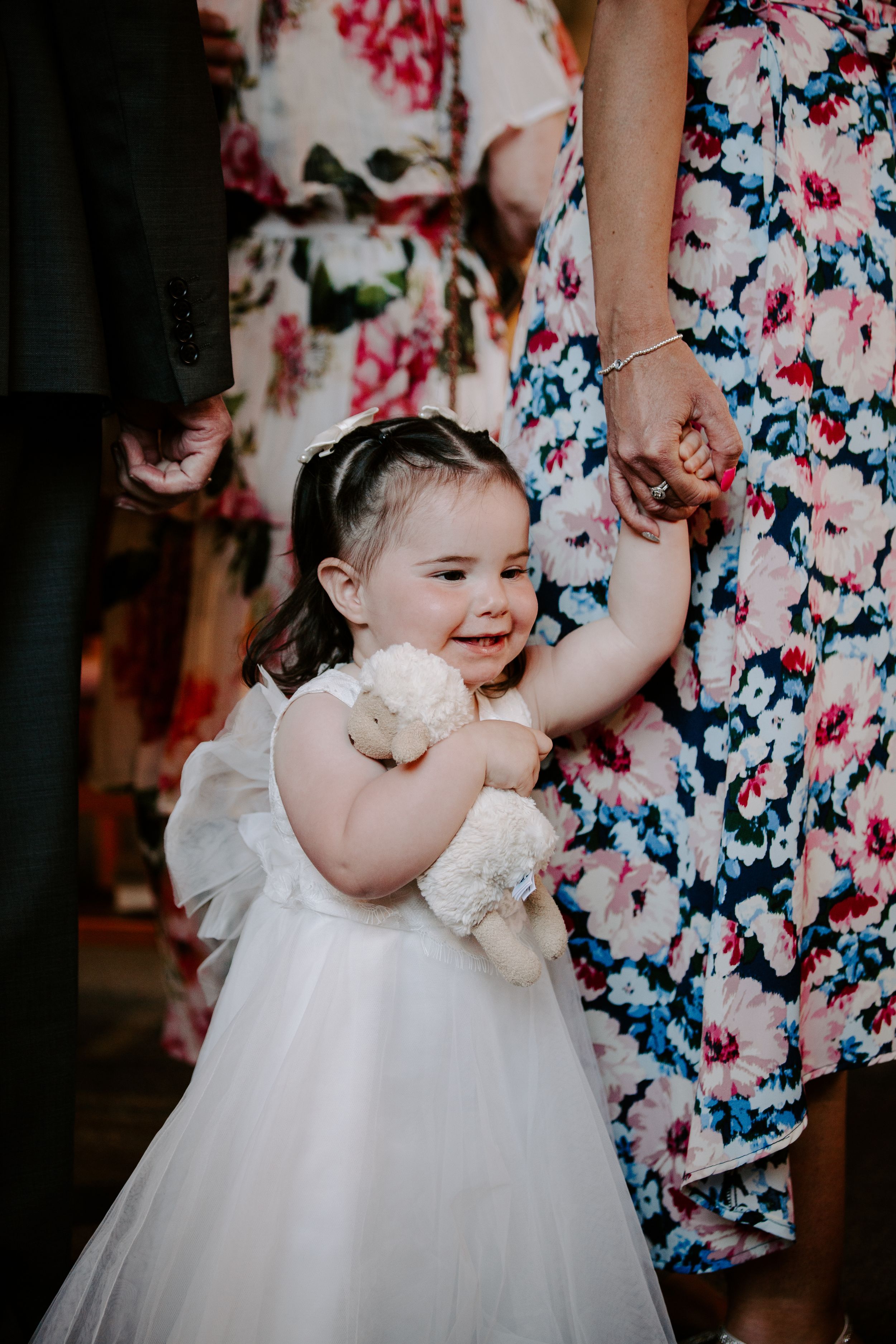 Adorable flower girl in white tutu dress holding stuffed animal, smiling joyfully at a wedding ceremony.