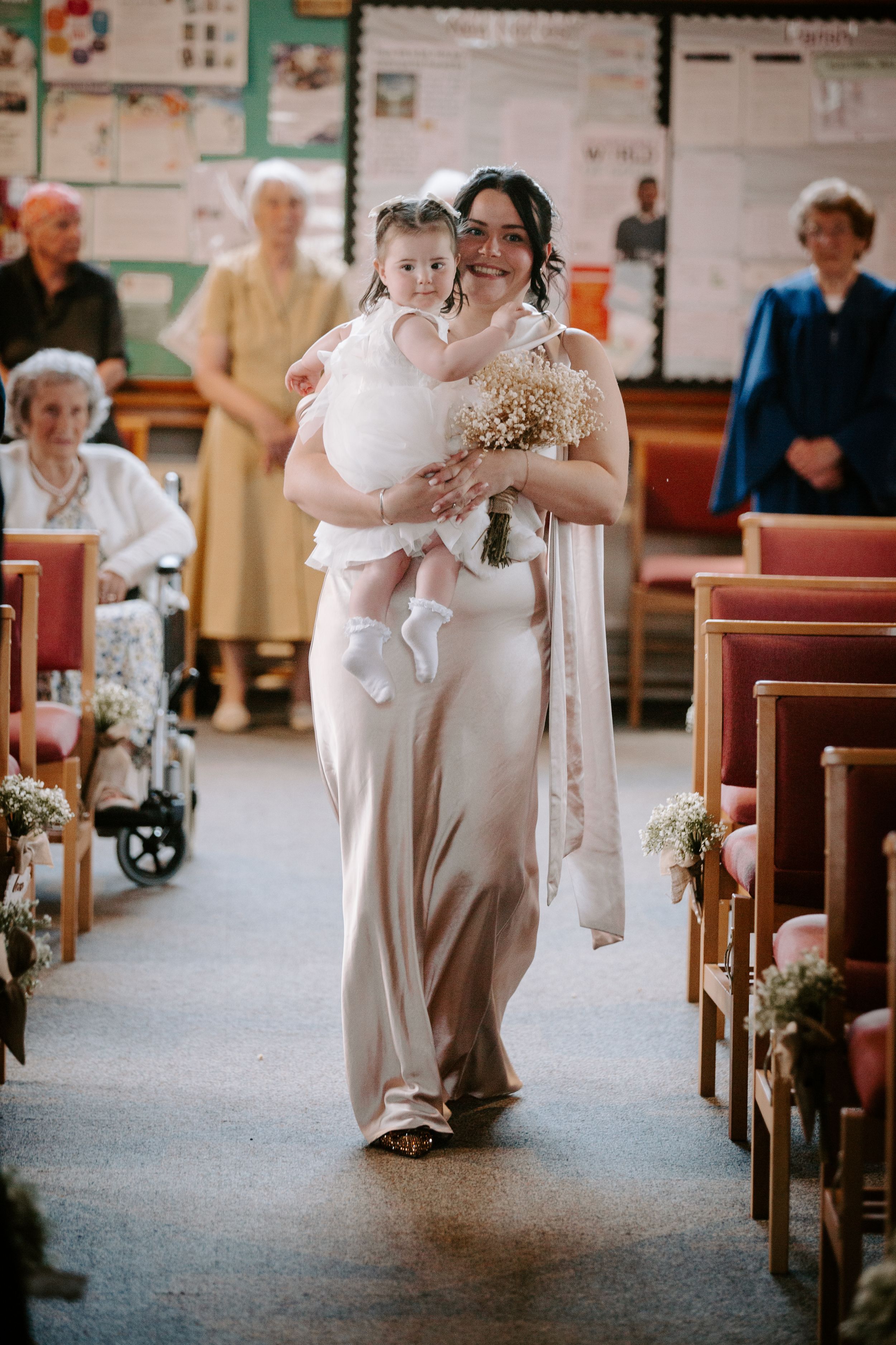 Bridesmaid in champagne gown carries flower girl down church aisle at wedding ceremony.