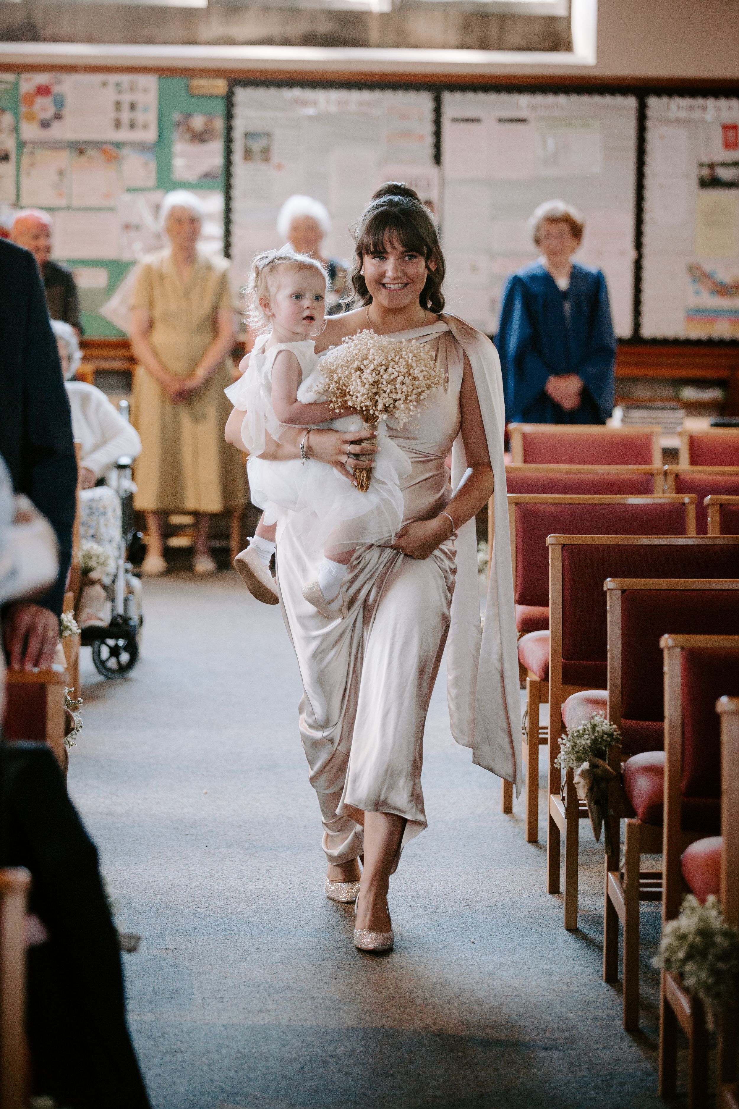 Bridesmaid in champagne satin dress carries flower girl down church aisle holding white baby's breath bouquet.