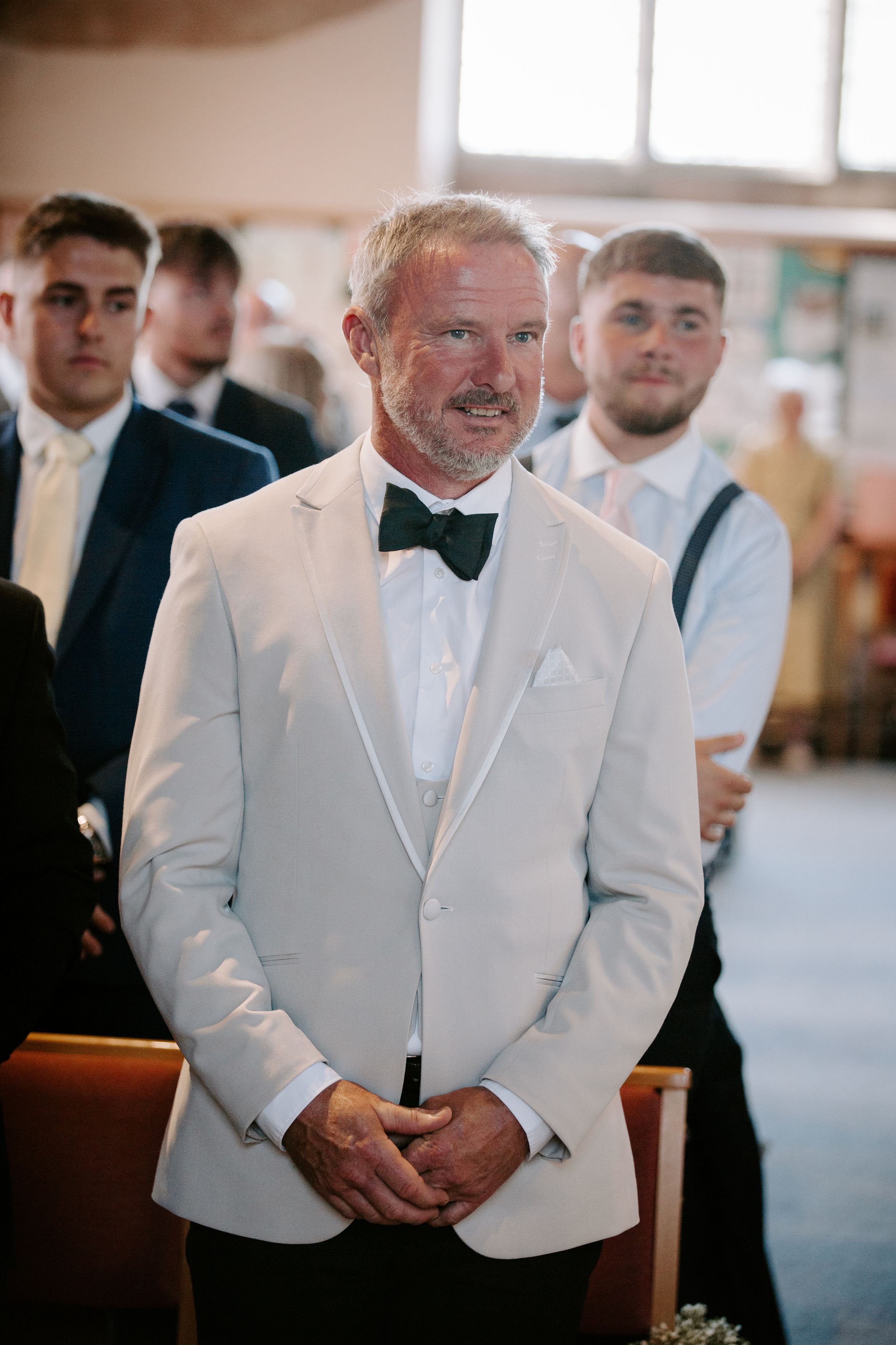 Groom in white tuxedo jacket and black bow tie stands at church altar, smiling with clasped hands.