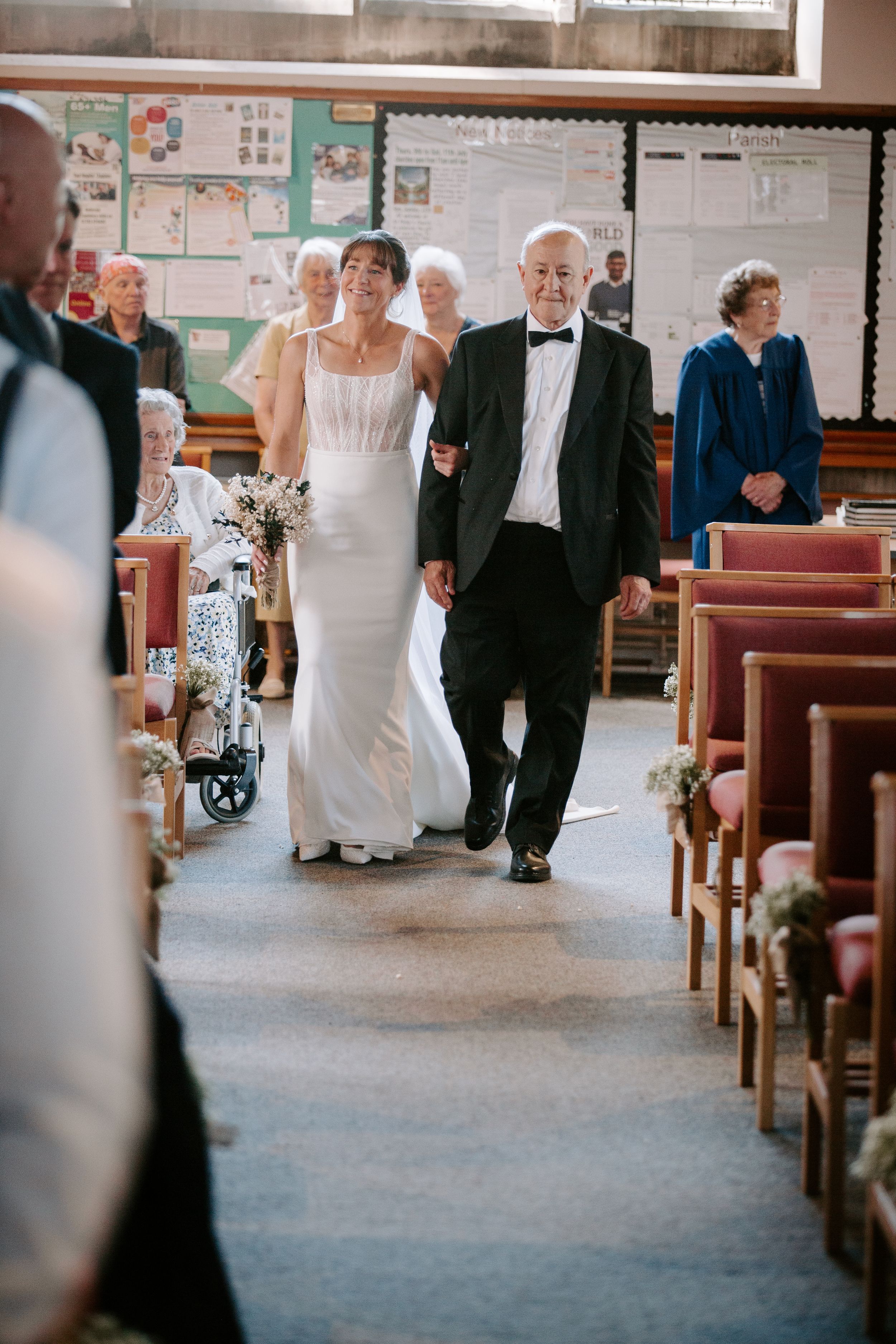 Bride in white gown walks down church aisle with elderly man in tuxedo during wedding ceremony.