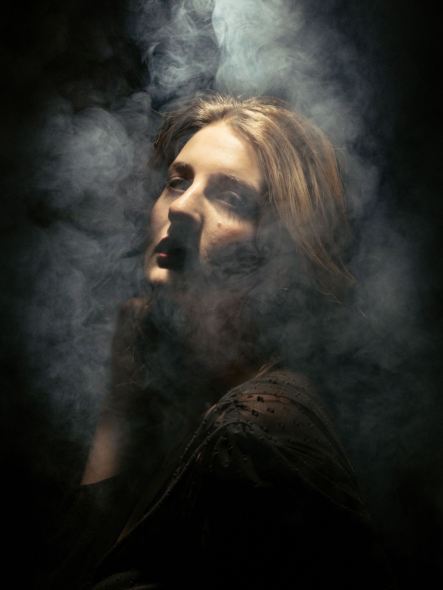 Dramatic portrait of a person with long hair surrounded by swirling smoke against a dark background.