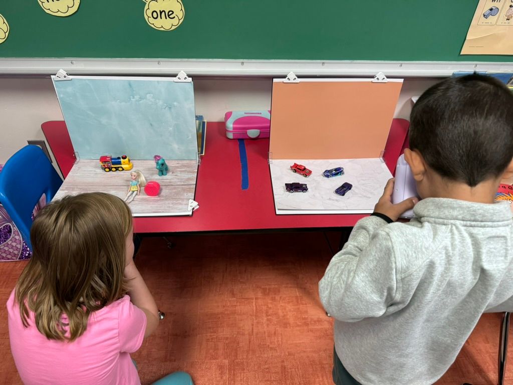 Two young children sit at a red table engaged in a classroom activity with colorful toys and paper boards.