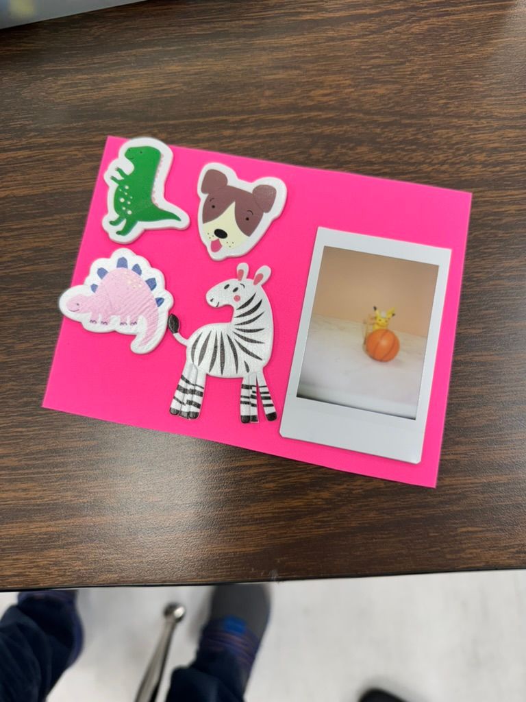 Pink card with animal stickers including zebra, dinosaurs, and dog, alongside a Polaroid photo on a wooden table.