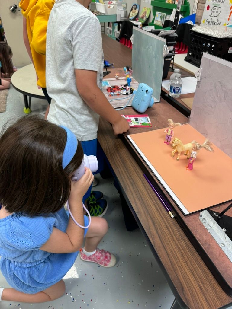 A young girl crouches to photograph toy figurines on orange paper in a colorful classroom setting.
