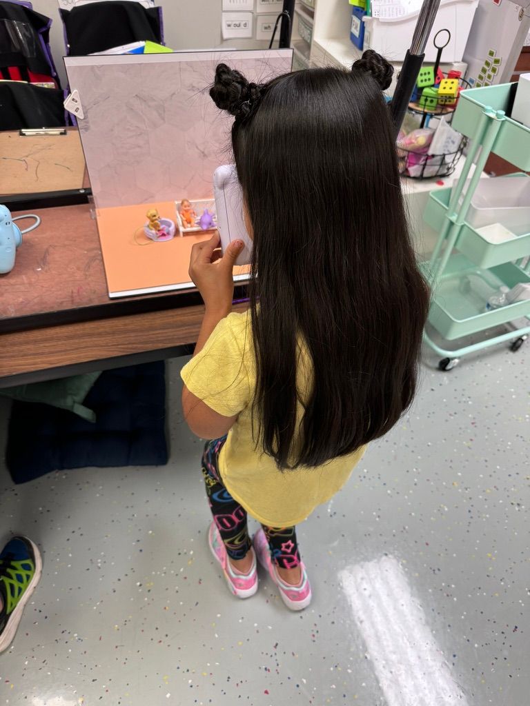 Young girl with long dark hair in pigtails wearing yellow shirt and colorful sneakers working at a classroom desk.