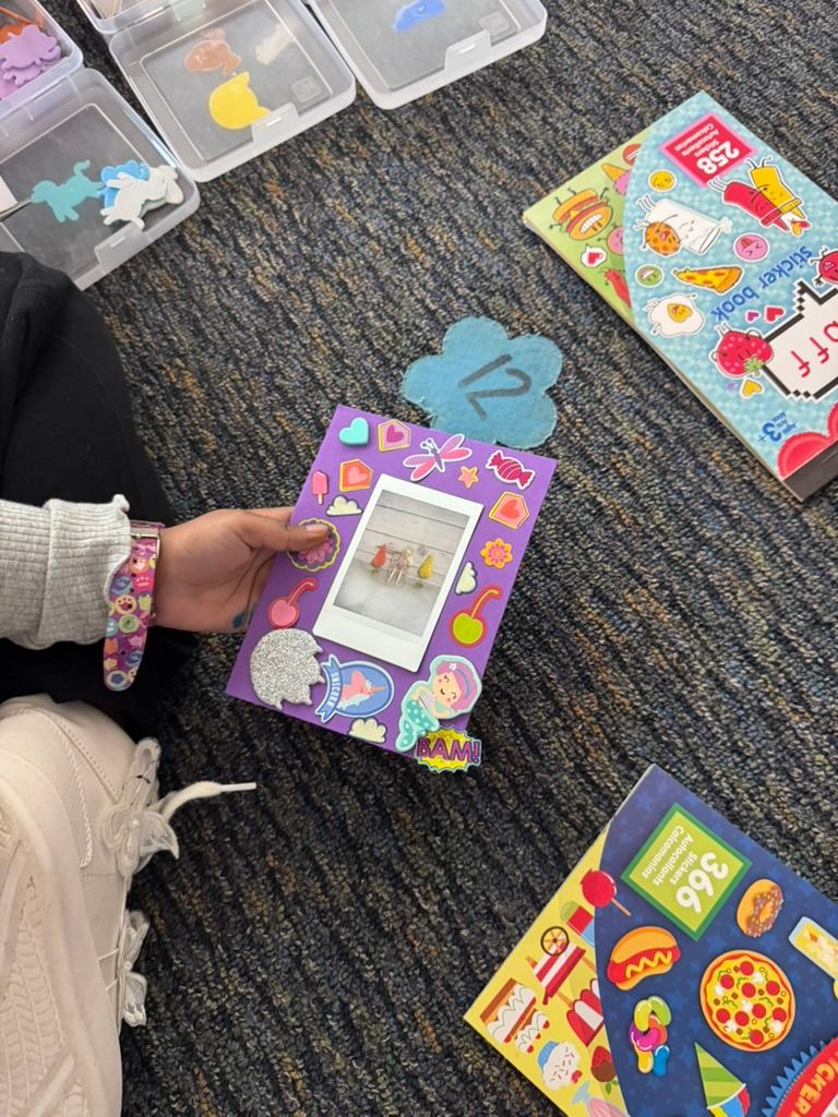 A child decorates a colorful sticker-covered photo frame on a carpeted floor surrounded by craft supplies and sticker books.