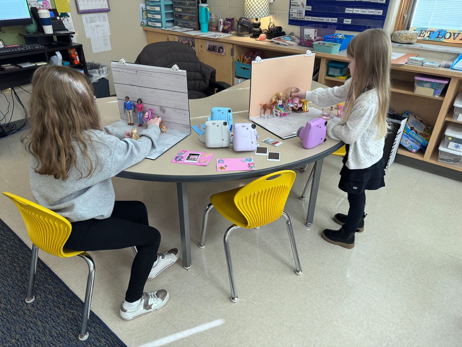 Two young girls work on creative projects at a kidney-shaped classroom table with yellow chairs in an elementary school.