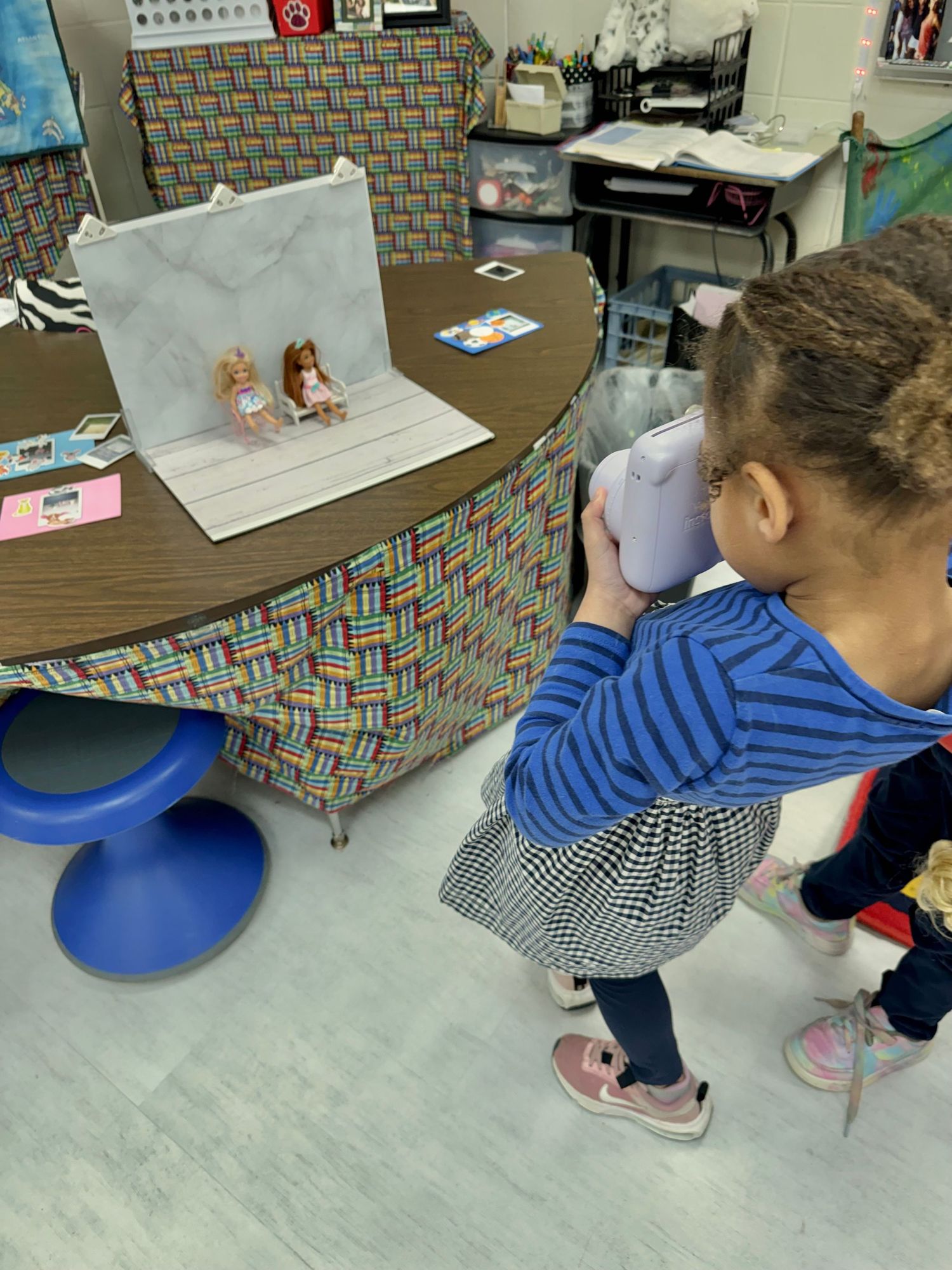 Young girl photographs dolls on a marble backdrop in a colorful classroom photography setup.