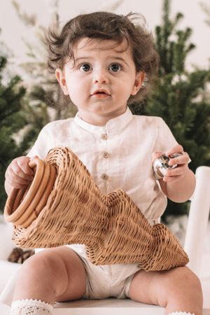 Small child in white outfit sits with wicker cornucopia and evergreen branches in background.