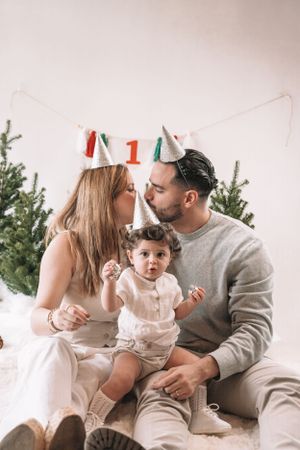 Family celebrates first birthday with decorative banner and evergreen trees in cozy indoor setting.