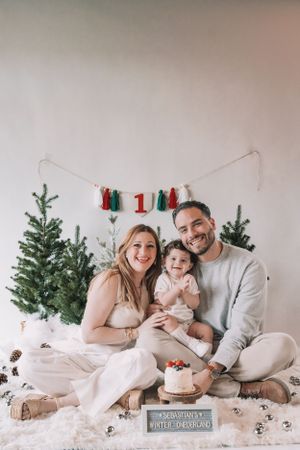 A festive family holiday photo with Christmas trees and decorative garland against a white wall.