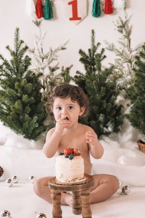 A small child sits with a decorated cake on a wooden stand surrounded by Christmas trees and holiday decor.