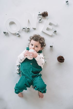 A toddler in green overalls lies on white backdrop with 'ONE' text and silver ornaments.