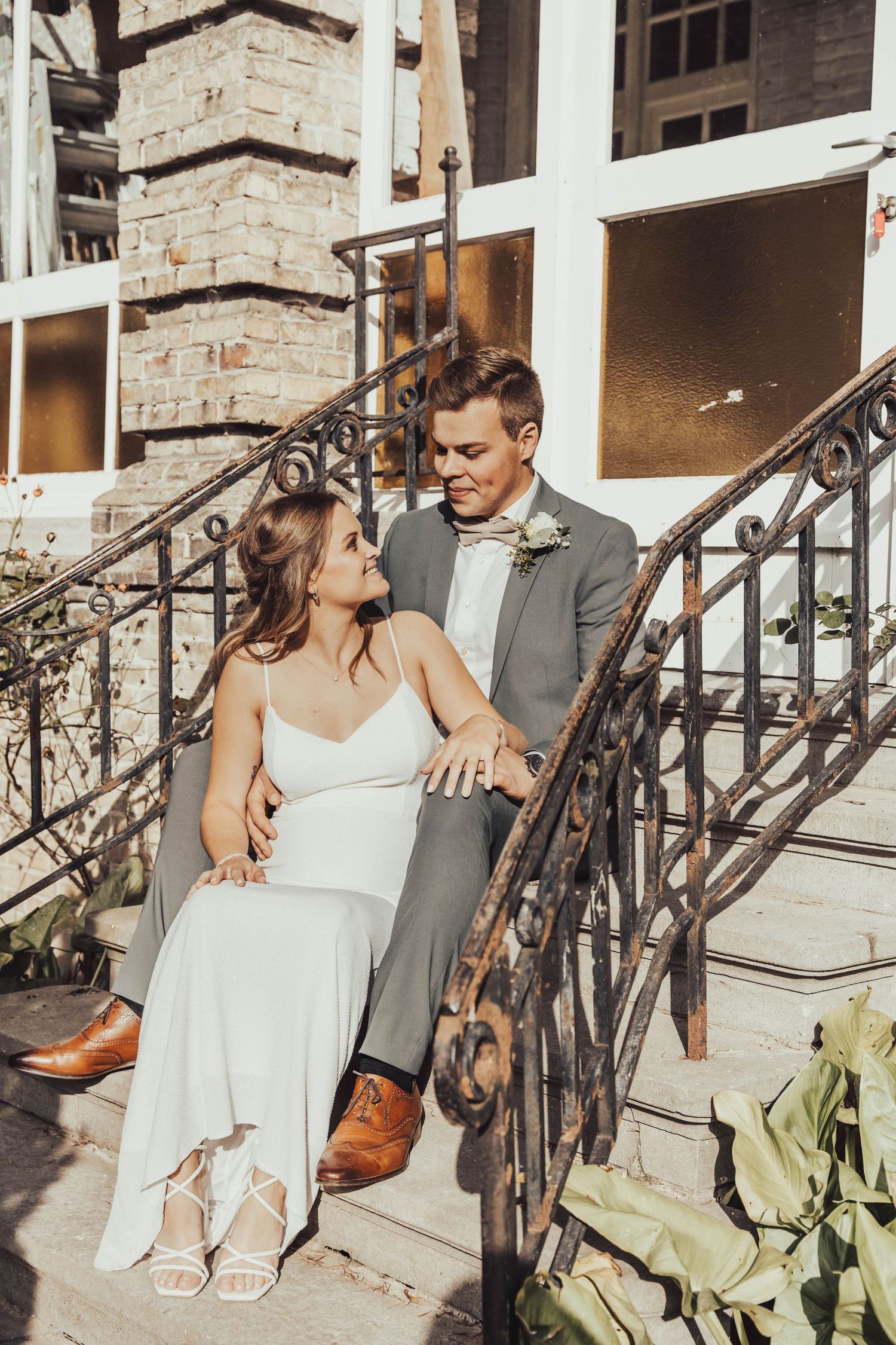 A couple sits together on outdoor stone steps, she in white and he in grey, sharing a candid moment in the sunlight.