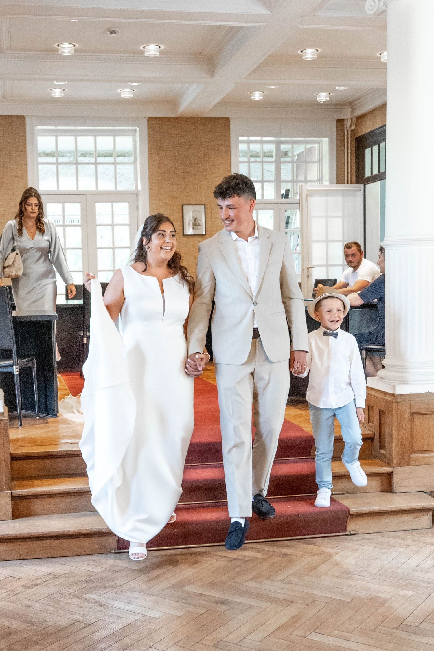 A wedding party descends red carpeted steps in an elegant room with coffered ceilings and large windows.