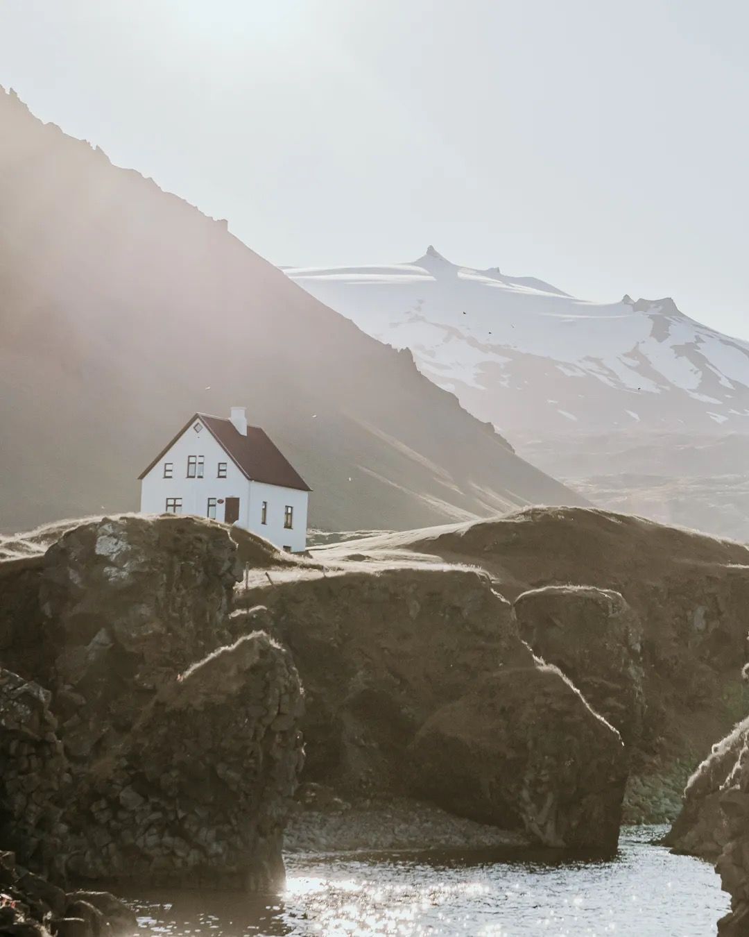 Lonely white house perched by cliffs with snowy mountains behind it.
