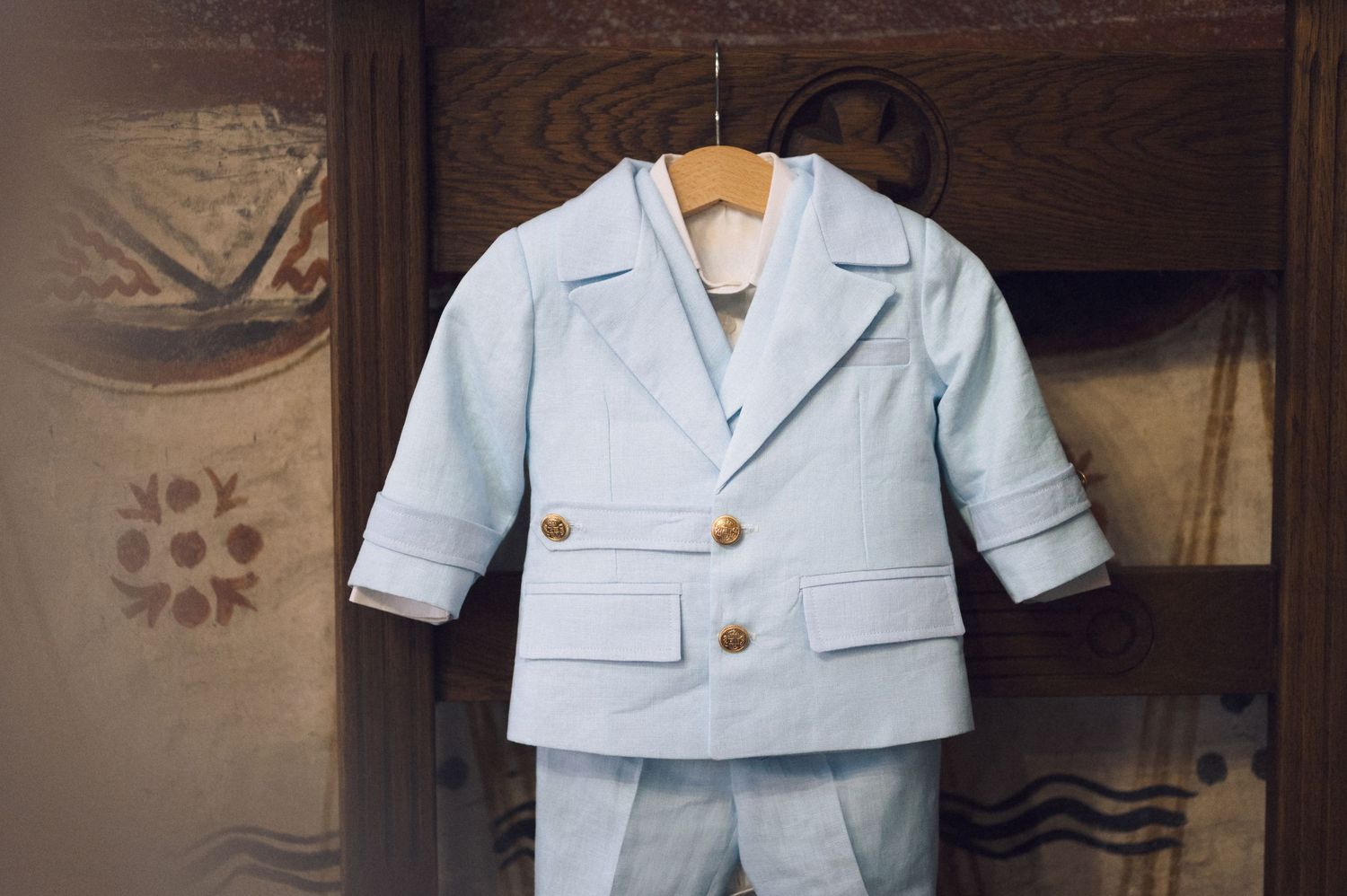 Light blue vintage baby boy suit with notched lapels and rolled cuffs hangs on wooden hanger against dark background.