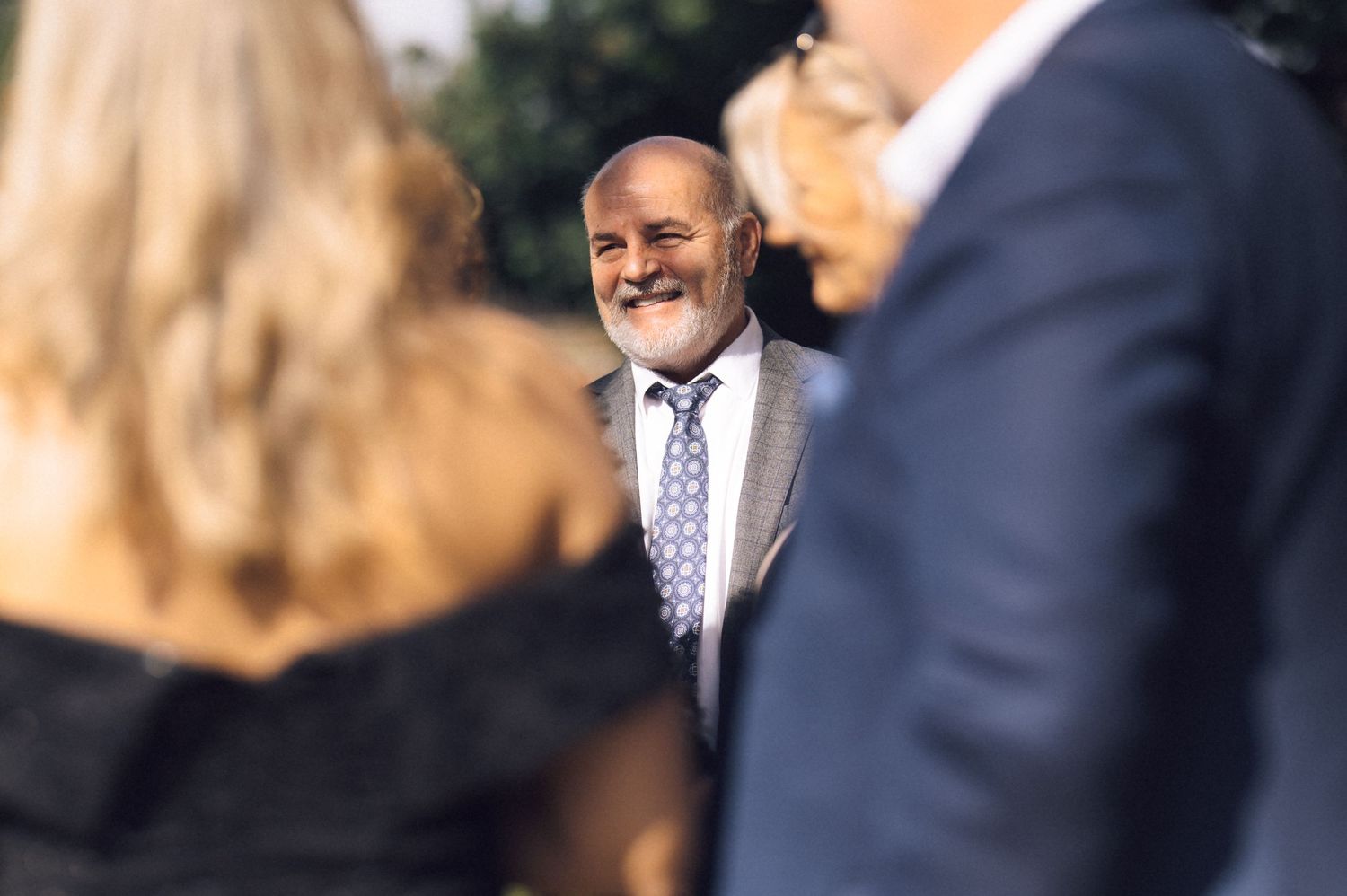 A person in a dark suit smiles warmly while standing outdoors in natural lighting.