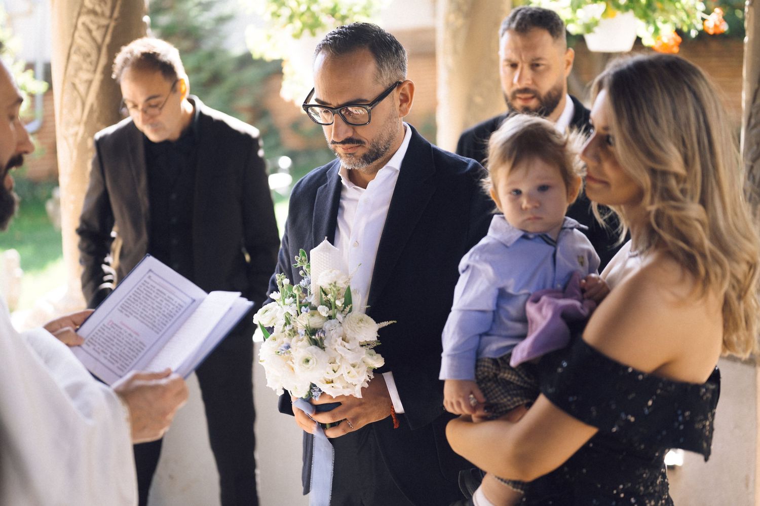 A small wedding ceremony takes place outdoors under a wooden pergola with guests gathered for an intimate celebration.