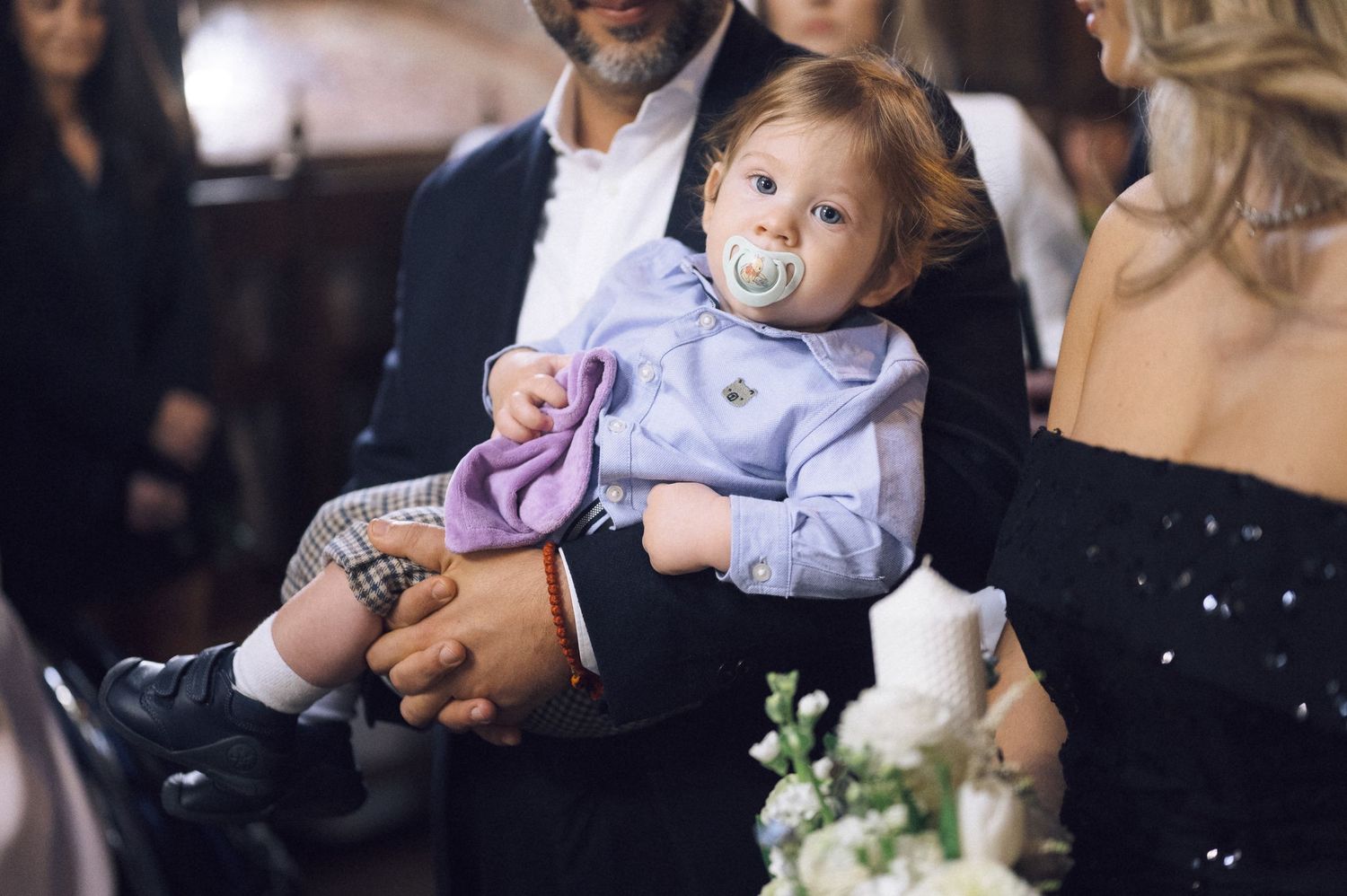A baby with a pacifier reaches for a white wedding cake while being held at a reception.