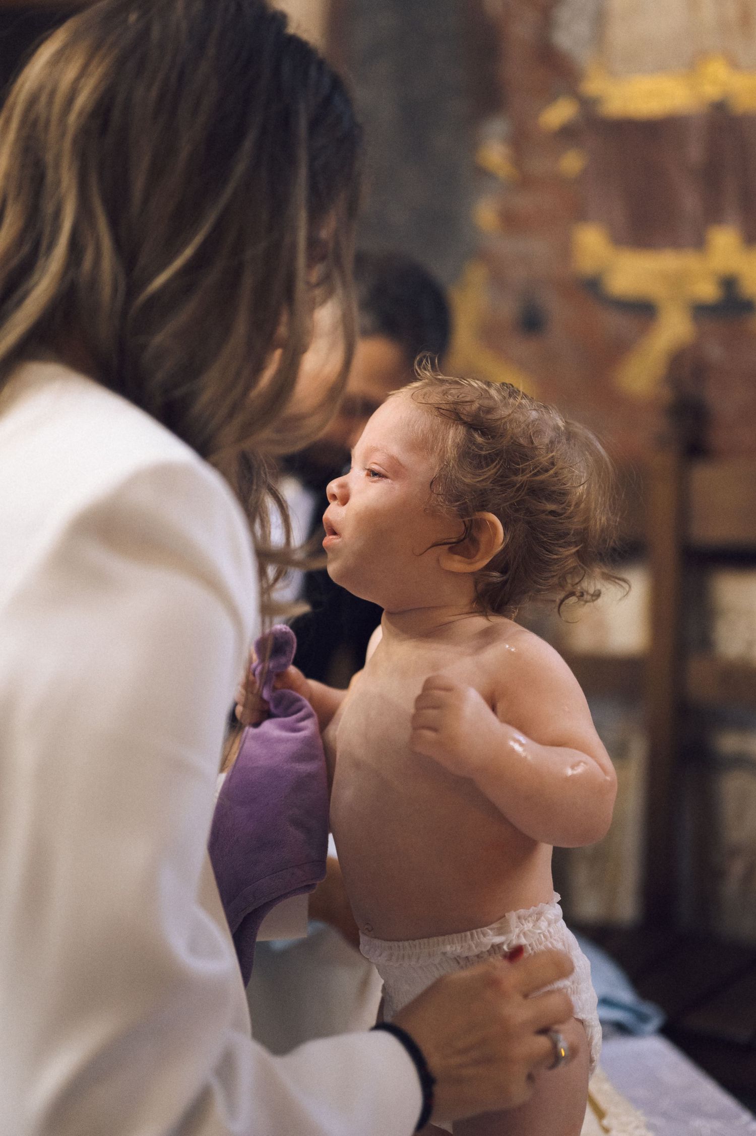 A tender moment between a caregiver in white and a shirtless baby with curly hair in a dimly lit setting.