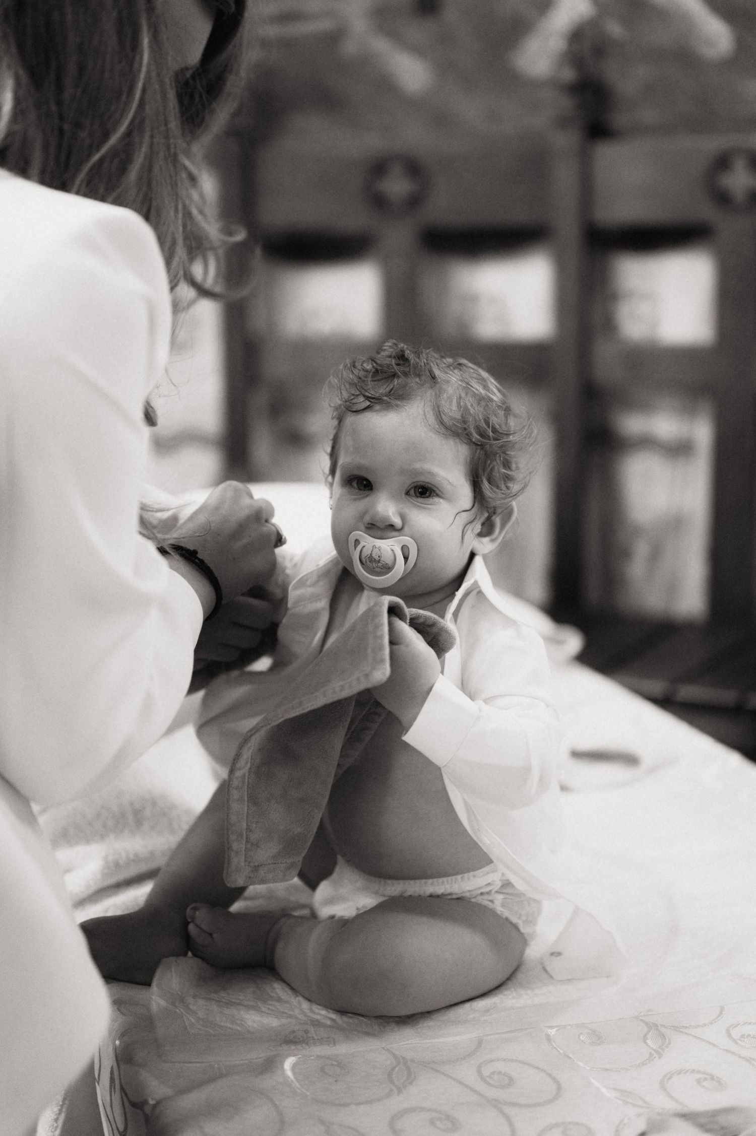 A black and white photograph shows a small child in white clothing sitting with a pacifier.