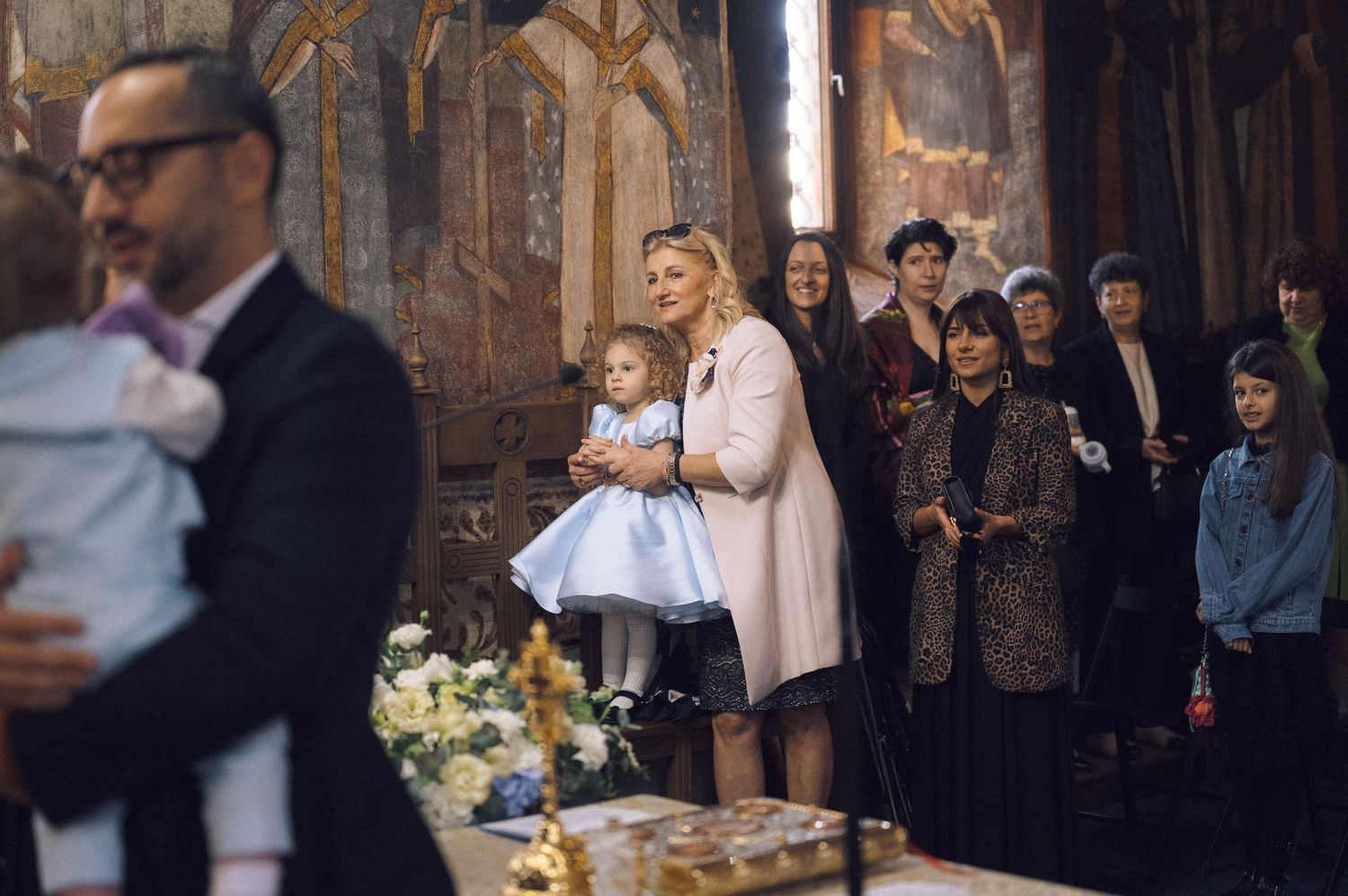 A group of people gather inside an ornate church during a baptism ceremony with decorative floral arrangements.