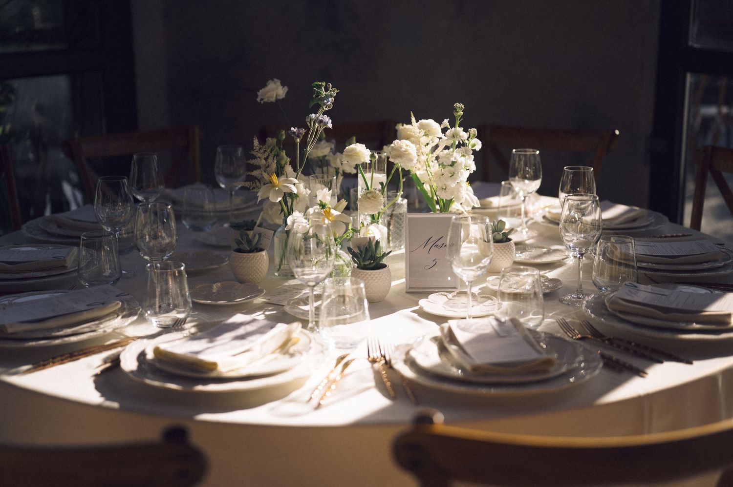 Elegant table settings with white plates and glassware illuminated by natural light streaming through windows.