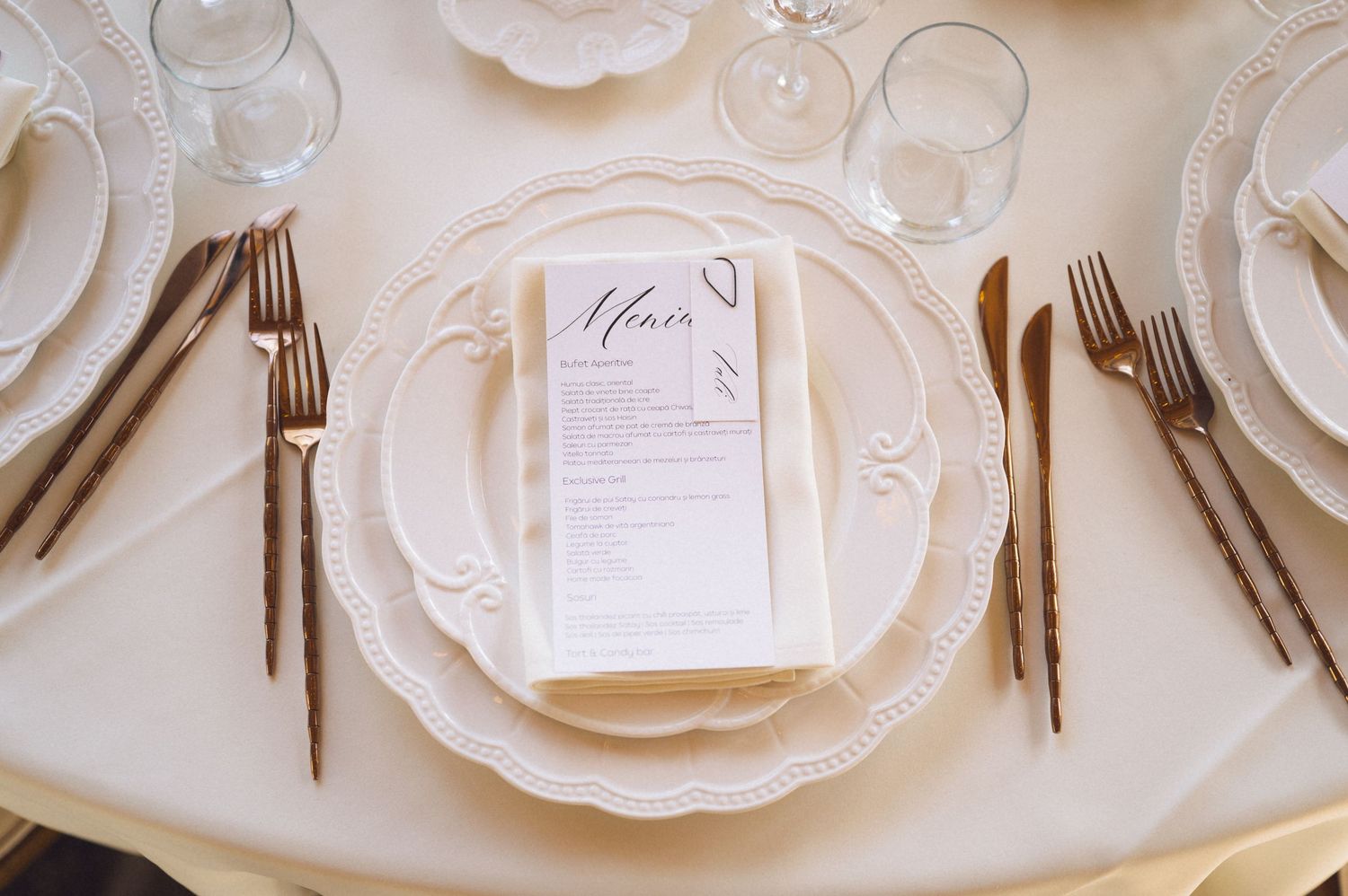 Elegant wedding place setting with white scalloped plates, vintage silverware, and a menu card on cream tablecloth.
