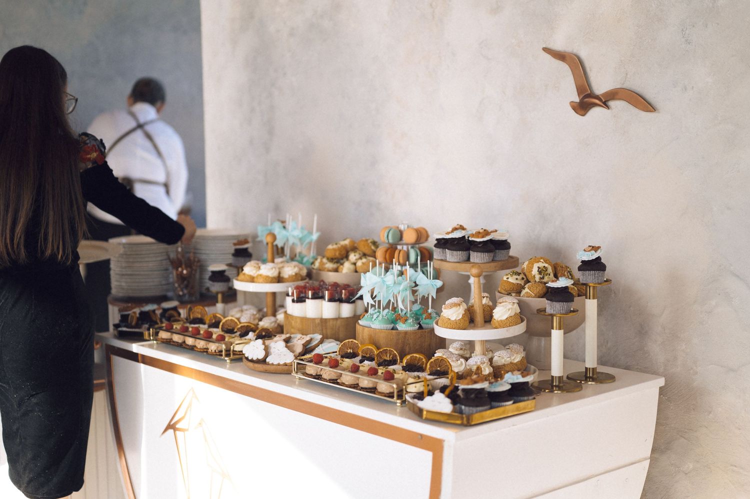 Elegant dessert table with cupcakes and pastries displayed on white stands against a neutral wall with decorative bird accents.