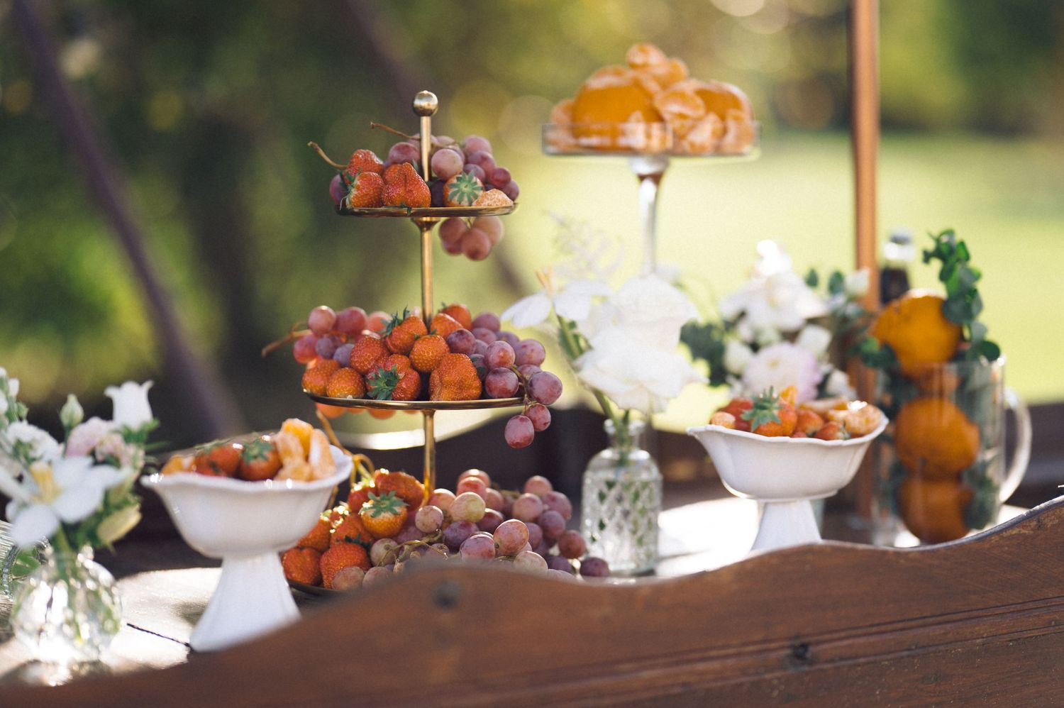Elegant dessert table display with tiered serving stands filled with pastries and fresh fruits surrounded by floral arrangements.