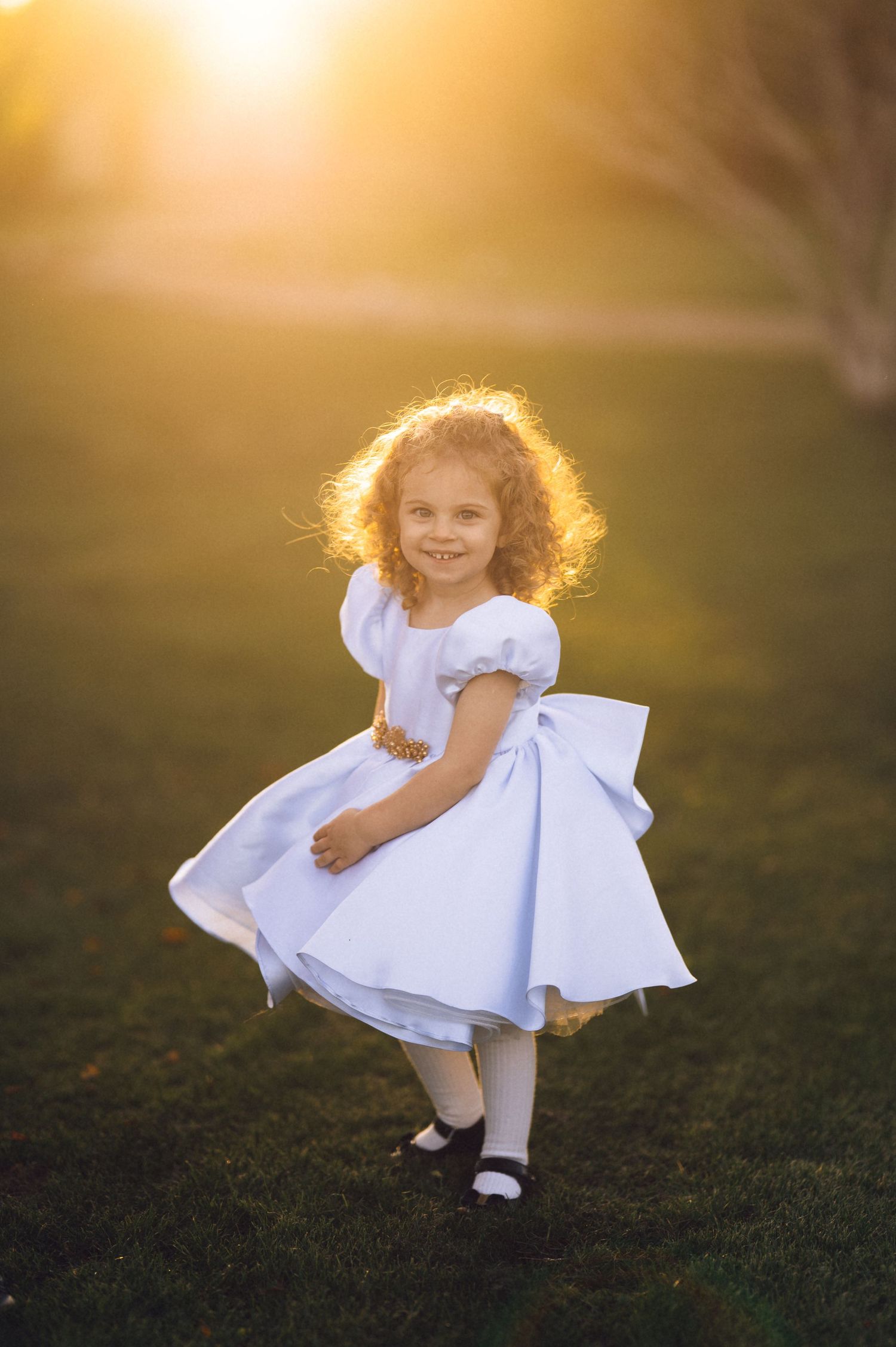 A young child in a white dress twirls playfully on green grass during golden hour sunlight.