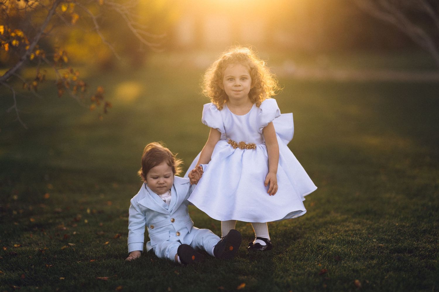 Children in white dresses sit together in a golden sunlit meadow during a dreamy autumn photoshoot.