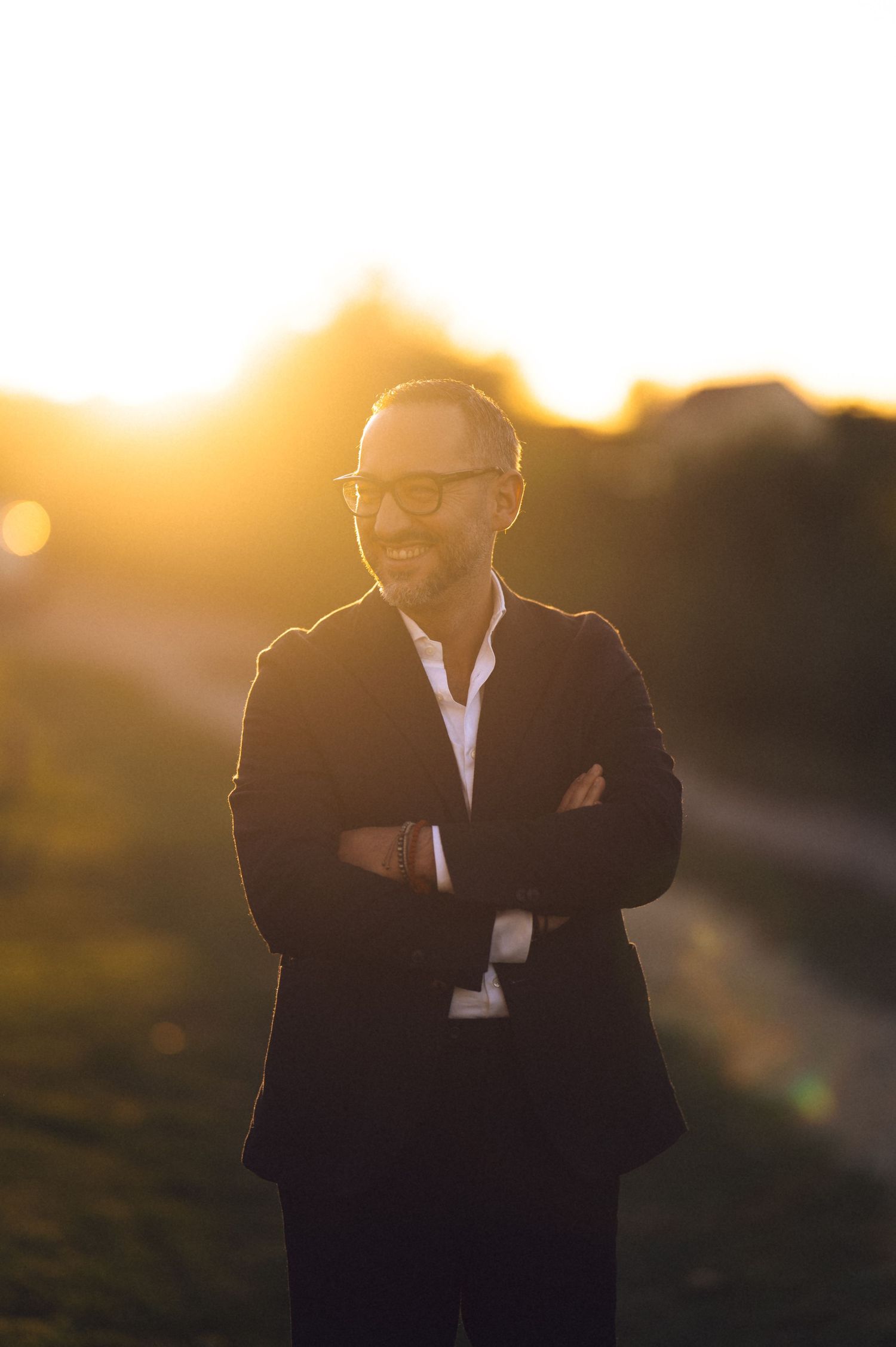 A person in a black suit smiles during golden hour sunset with warm glowing bokeh in the background.