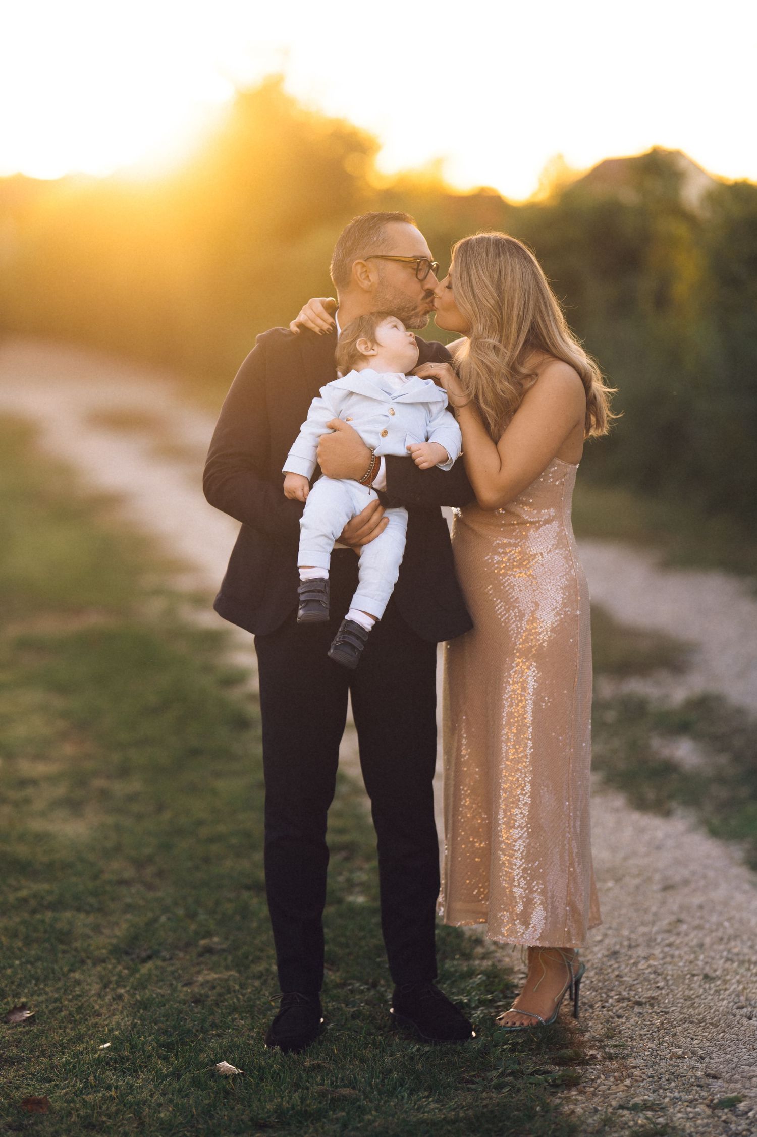 A couple shares an intimate moment with their baby at sunset while standing on a dirt path, the woman wearing a sparkly gold dress.