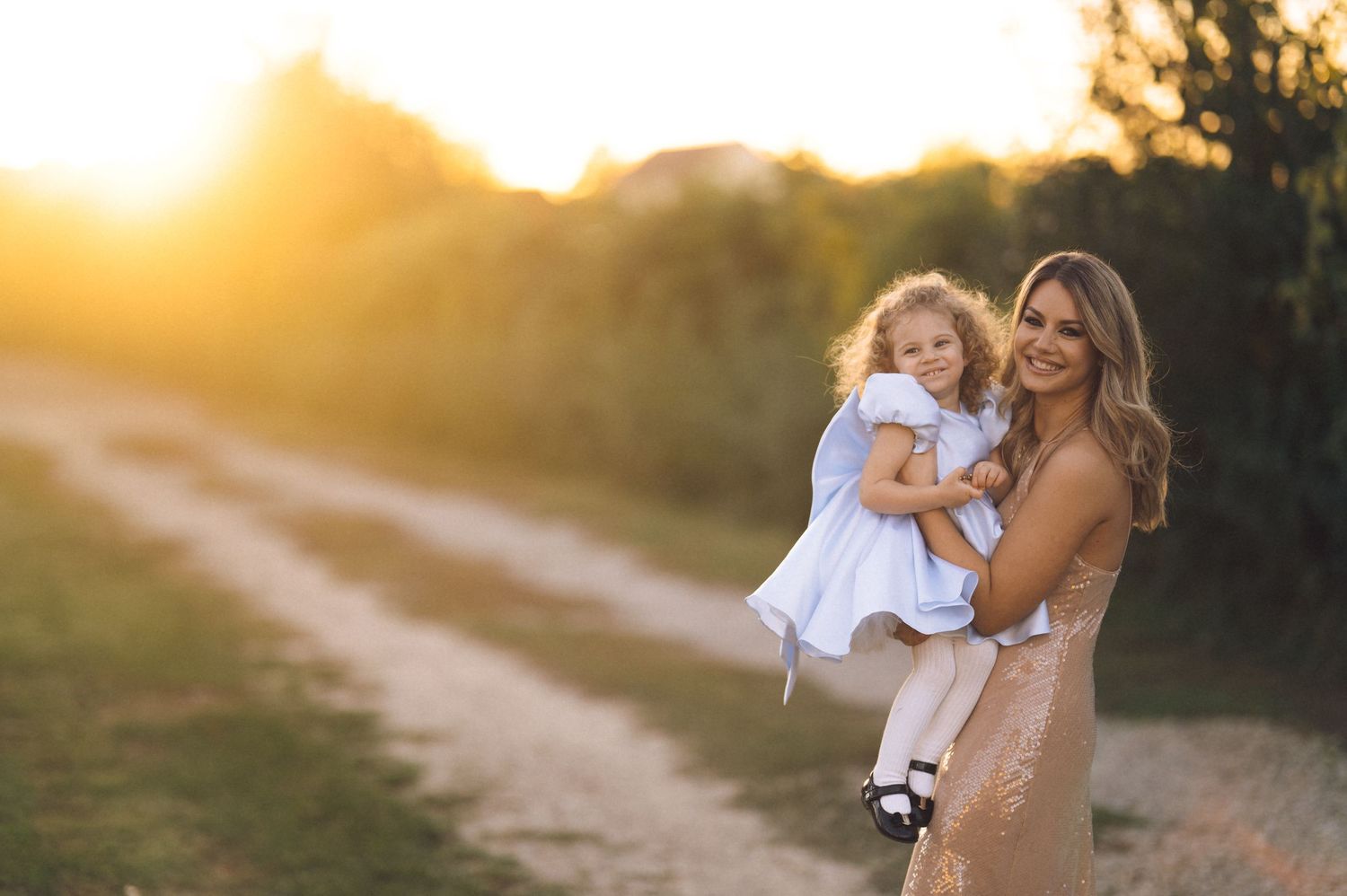 A mother and daughter share a joyful moment together while walking on a dirt path at golden hour sunset.