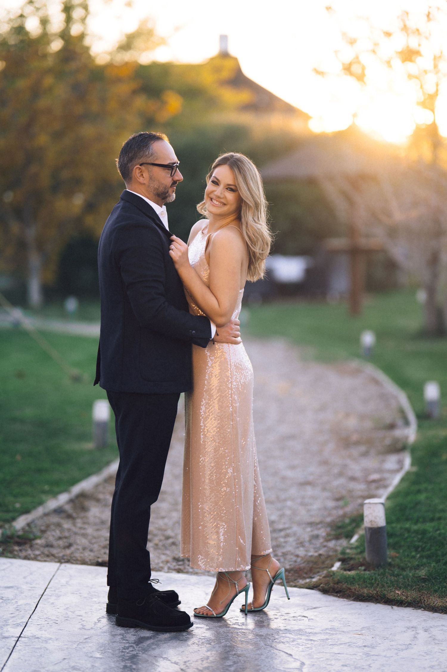 A couple shares a romantic moment at sunset on a path, with the person wearing a sparkly champagne-colored dress.
