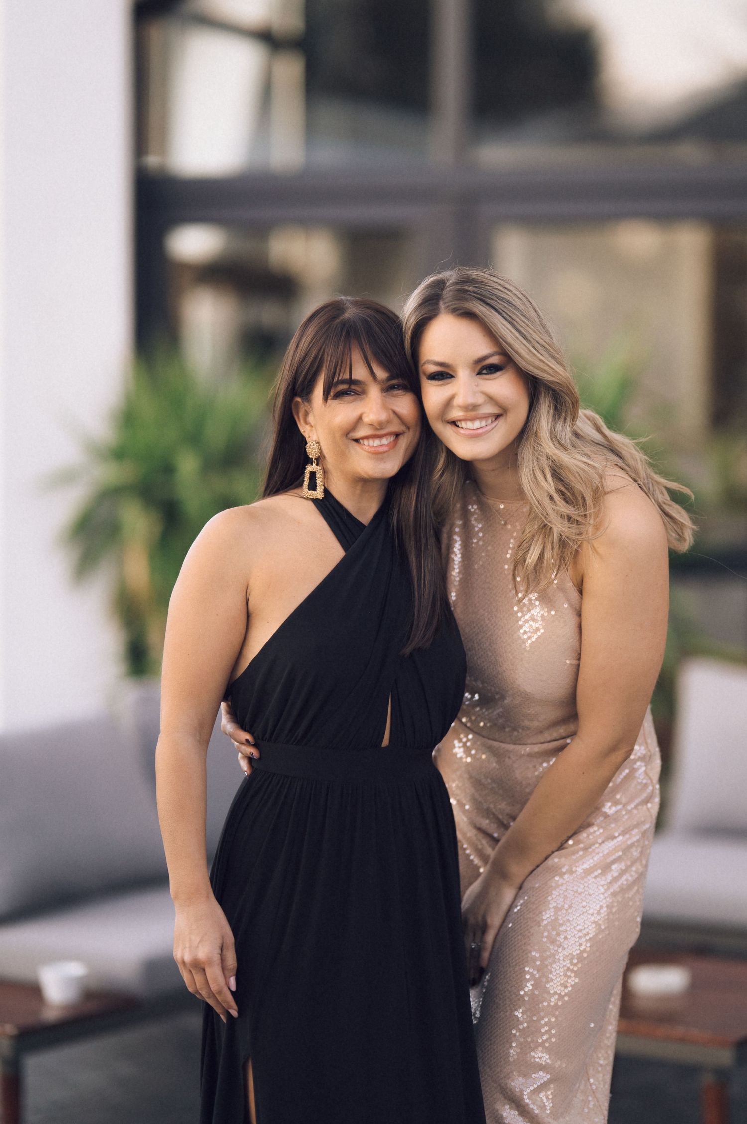 Two women pose together outdoors wearing contrasting black and sparkly champagne-colored evening dresses.