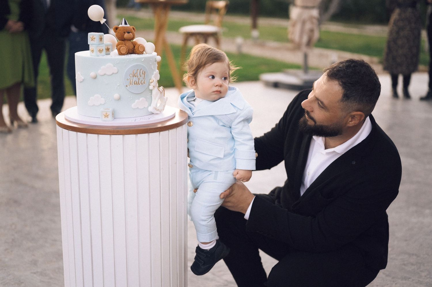 A baby in a white christening gown is held near a wooden baptismal font during a religious ceremony.