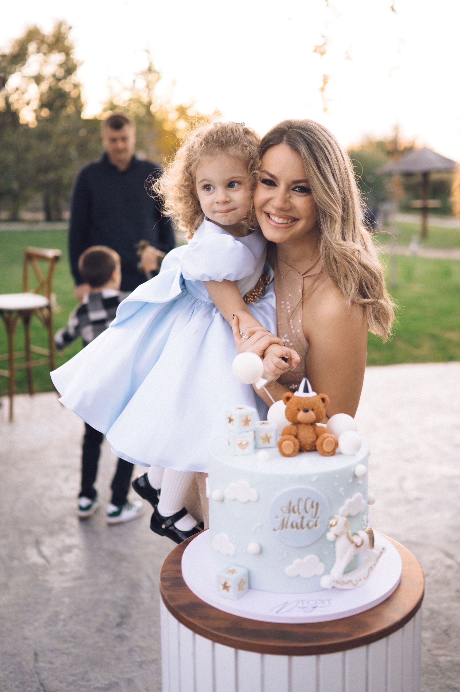 A series of baptism celebration photos featuring a white dress, teddy bear and decorative cake on a wooden table outdoors.