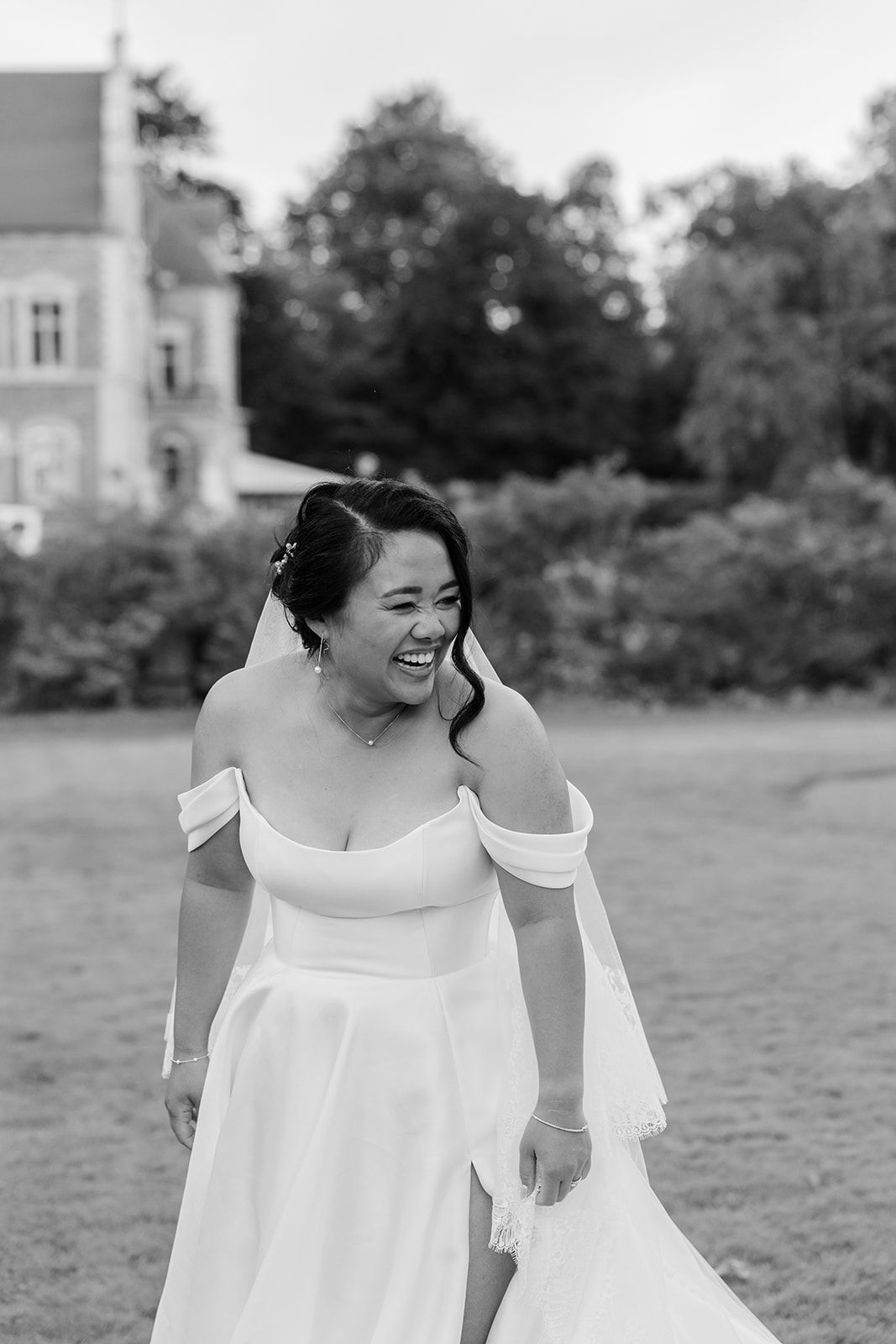 A person in a white off-shoulder dress smiles while looking down in an outdoor garden setting.
