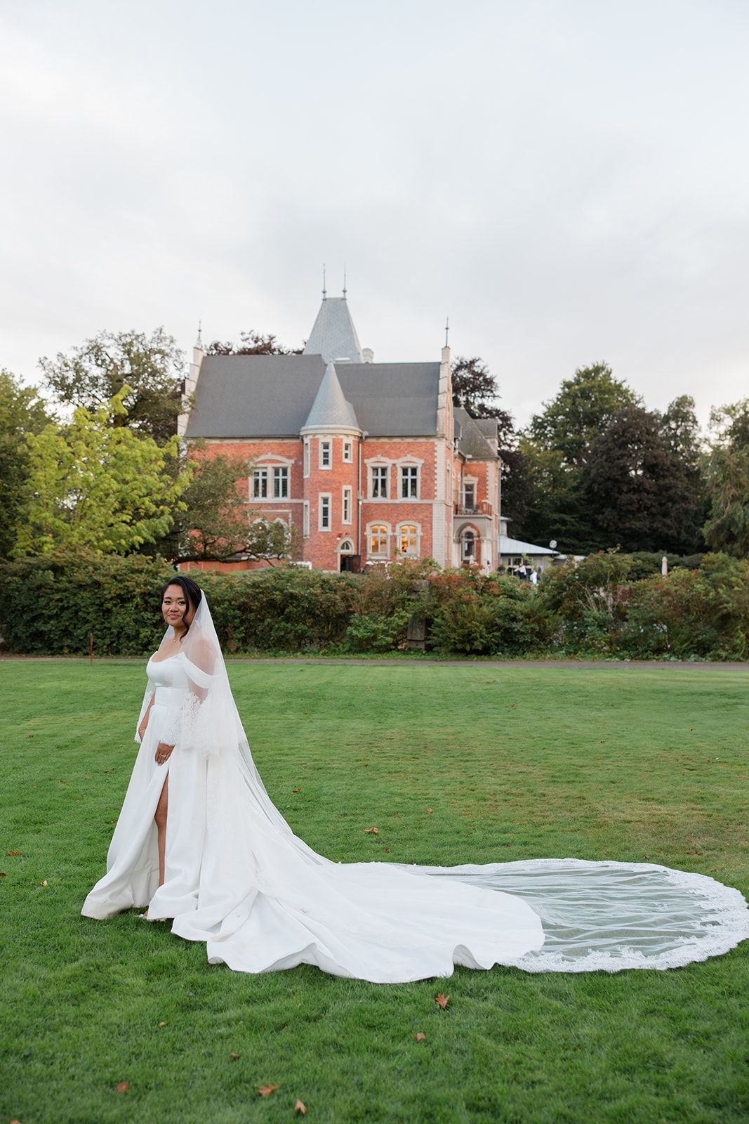 A bride in a flowing white gown with a dramatic cathedral-length veil poses on a green lawn with a brick mansion in the background.