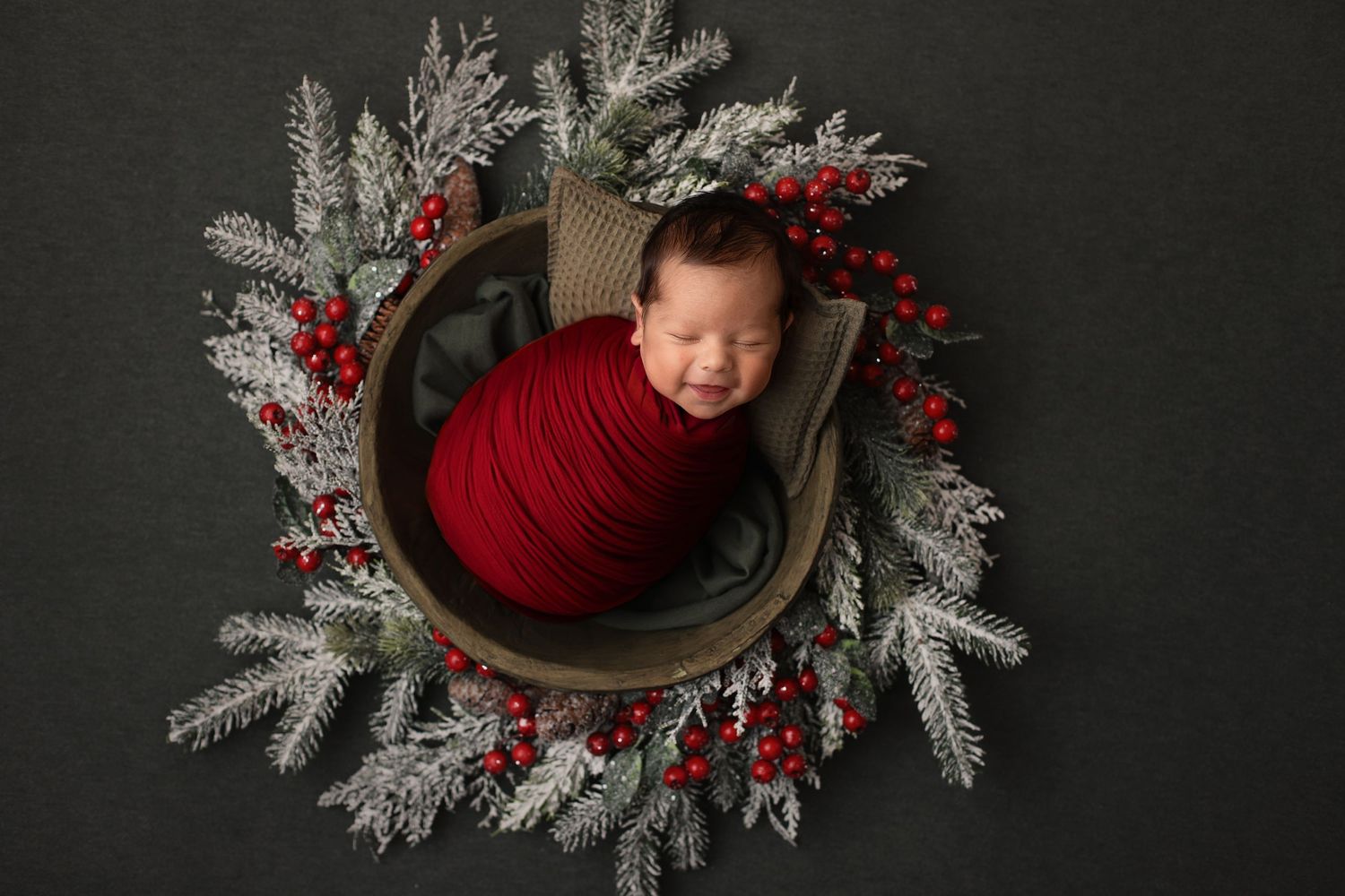 A sleeping baby wrapped in red fabric nestles in a wooden bowl surrounded by snowy branches and festive red berries - leander newborn photos
