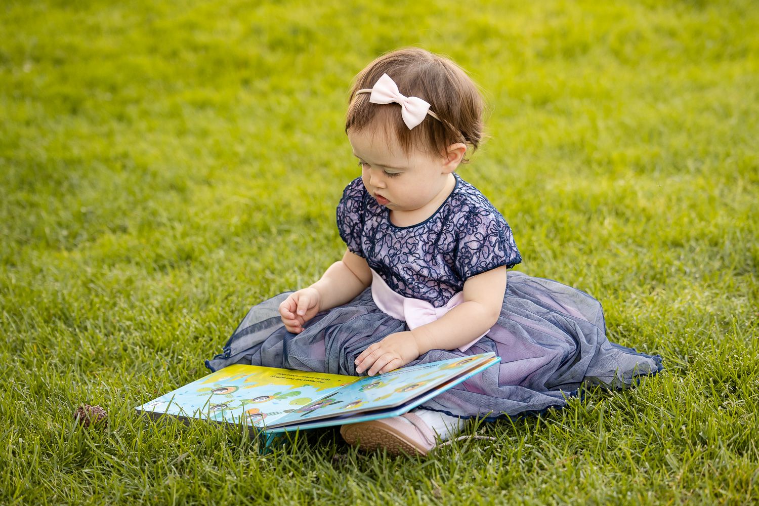 A young child in a dress sits on grass reading a picture book on a sunny day.